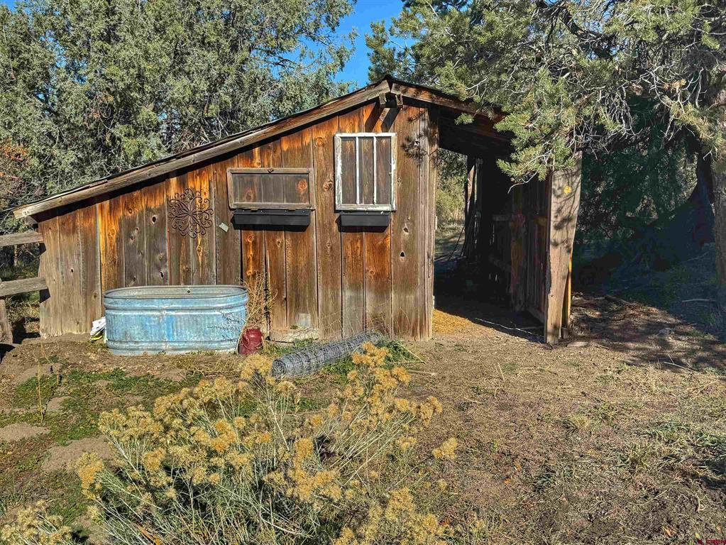 Weathered wooden outbuilding with a covered passage and galvanized trough among evergreen trees.
