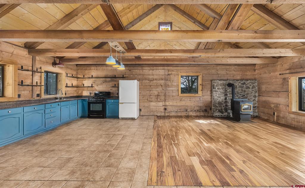 Rustic open kitchen and living area with blue cabinets, exposed timber beams, and a stone hearth with wood stove.
