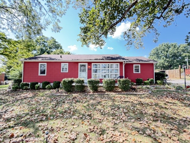 Red single-story house with white trim, front shrubs, and a leaf-covered lawn beneath mature trees.