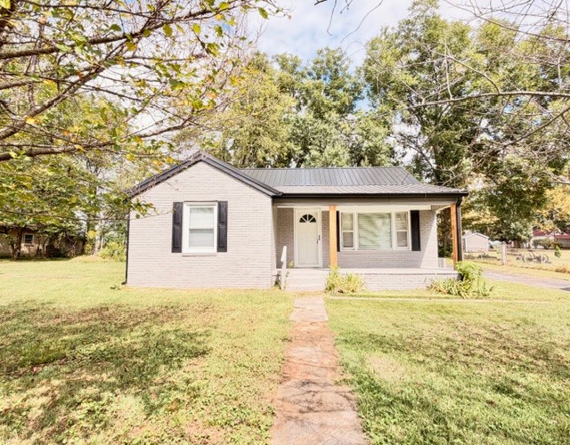 Single-story light-colored house with a dark metal roof and front walkway beneath mature trees.