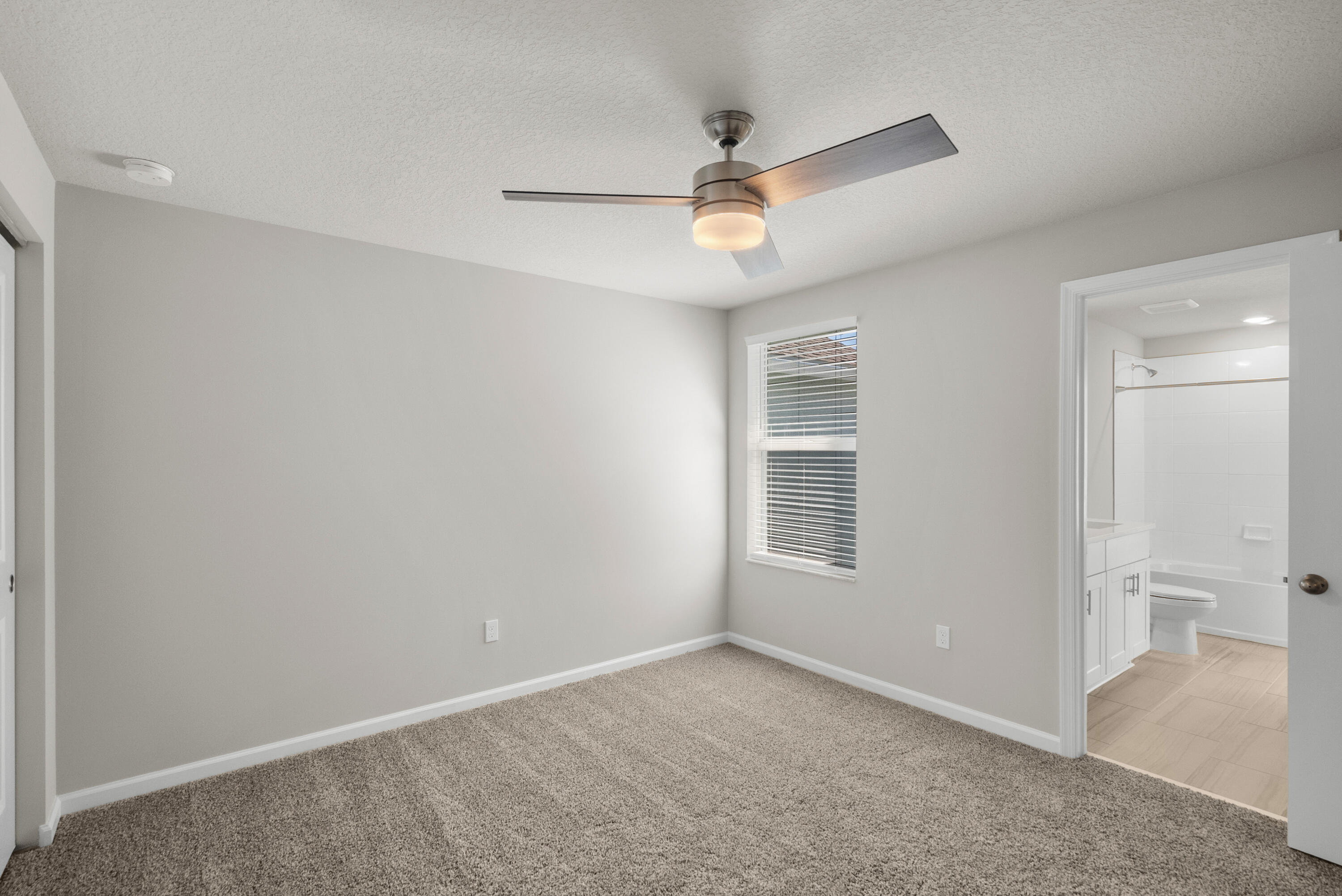 Empty carpeted bedroom with a ceiling fan and window; open doorway shows the adjoining bathroom.