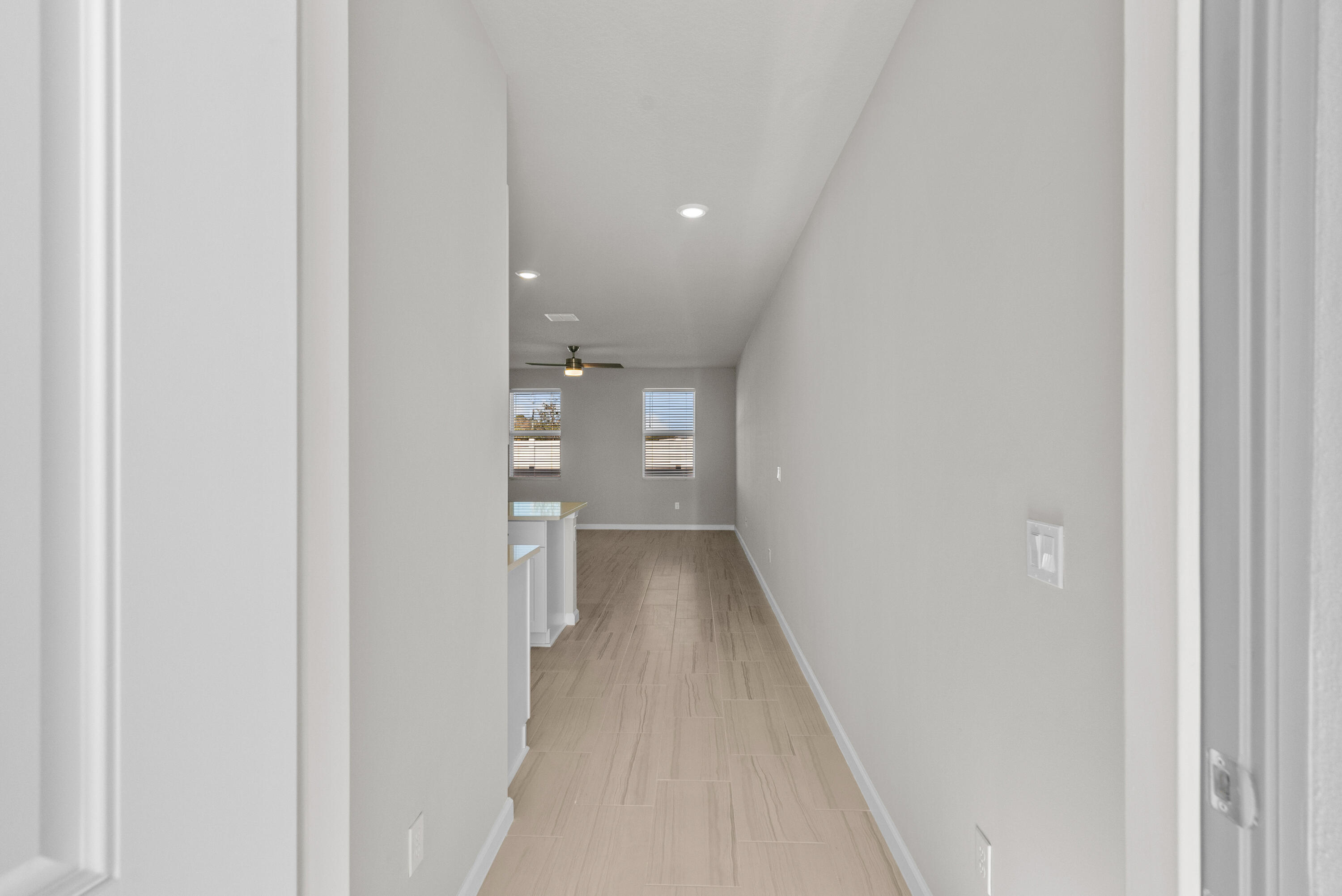 Empty hallway with light wood-look tile flooring, two far windows, and a partial kitchen counter.