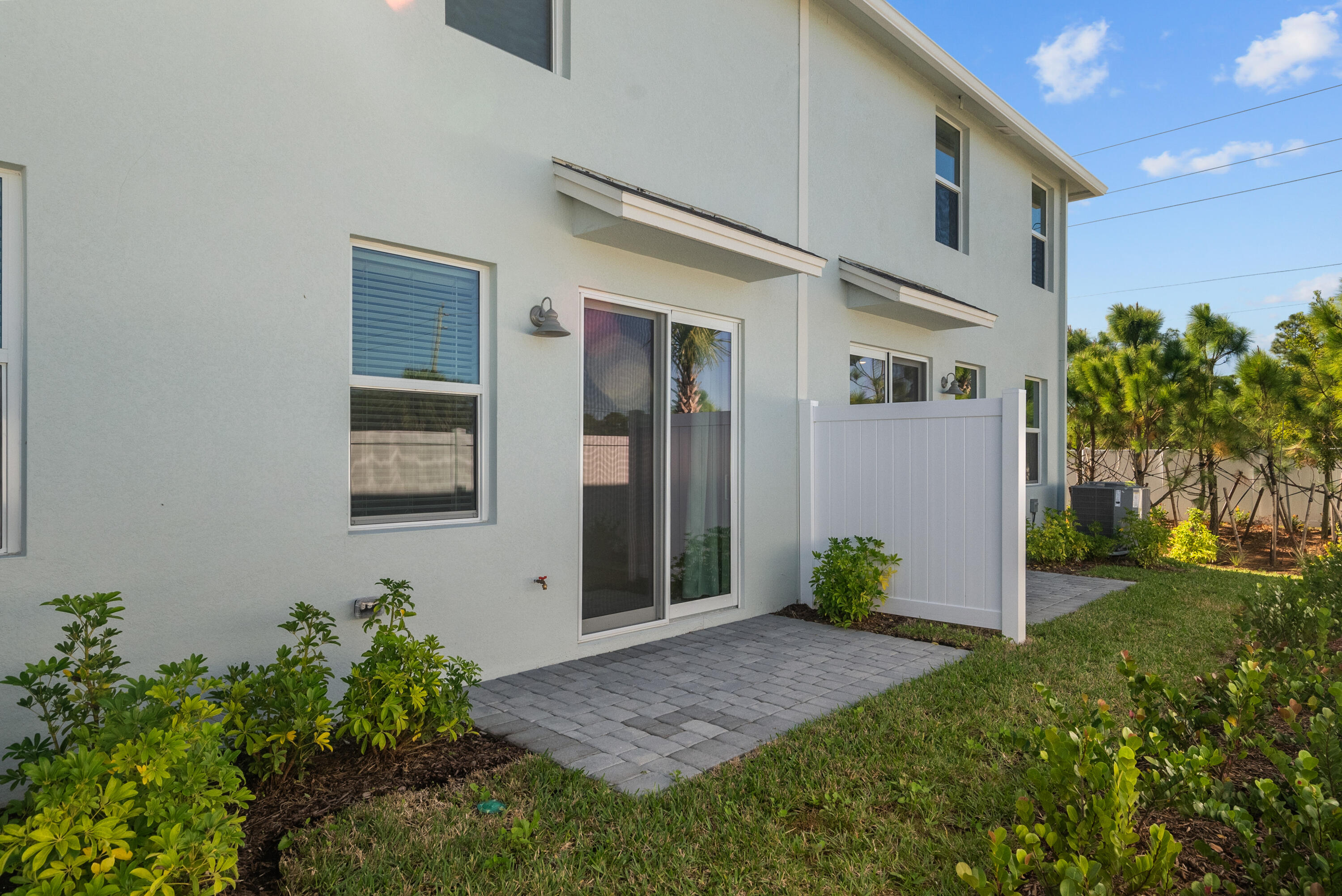 Two-story home with a gray paver patio, white privacy fence, and tropical landscaping.