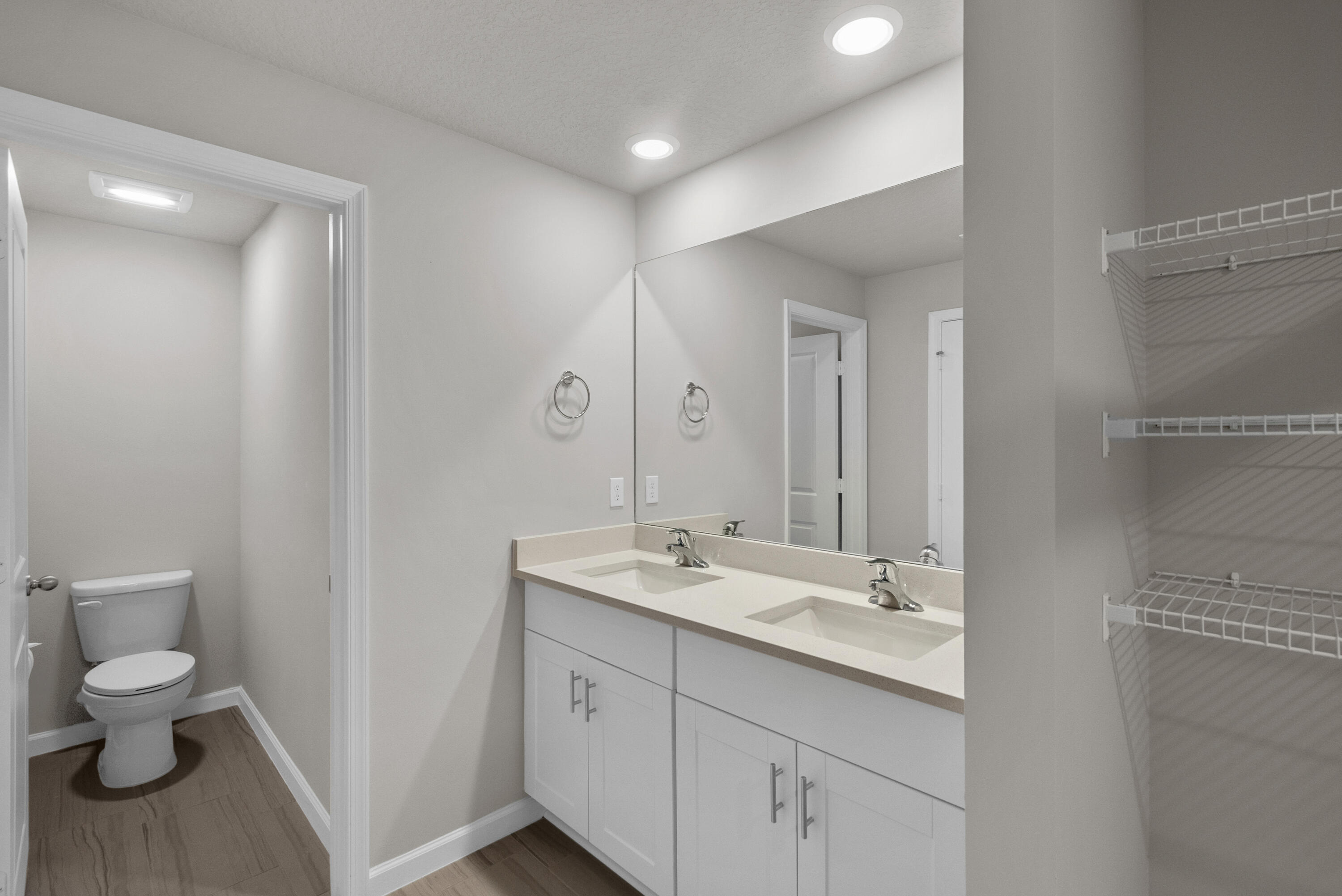 Bathroom with a double-sink vanity and mirror, adjoining toilet room, and open wire-shelf closet.