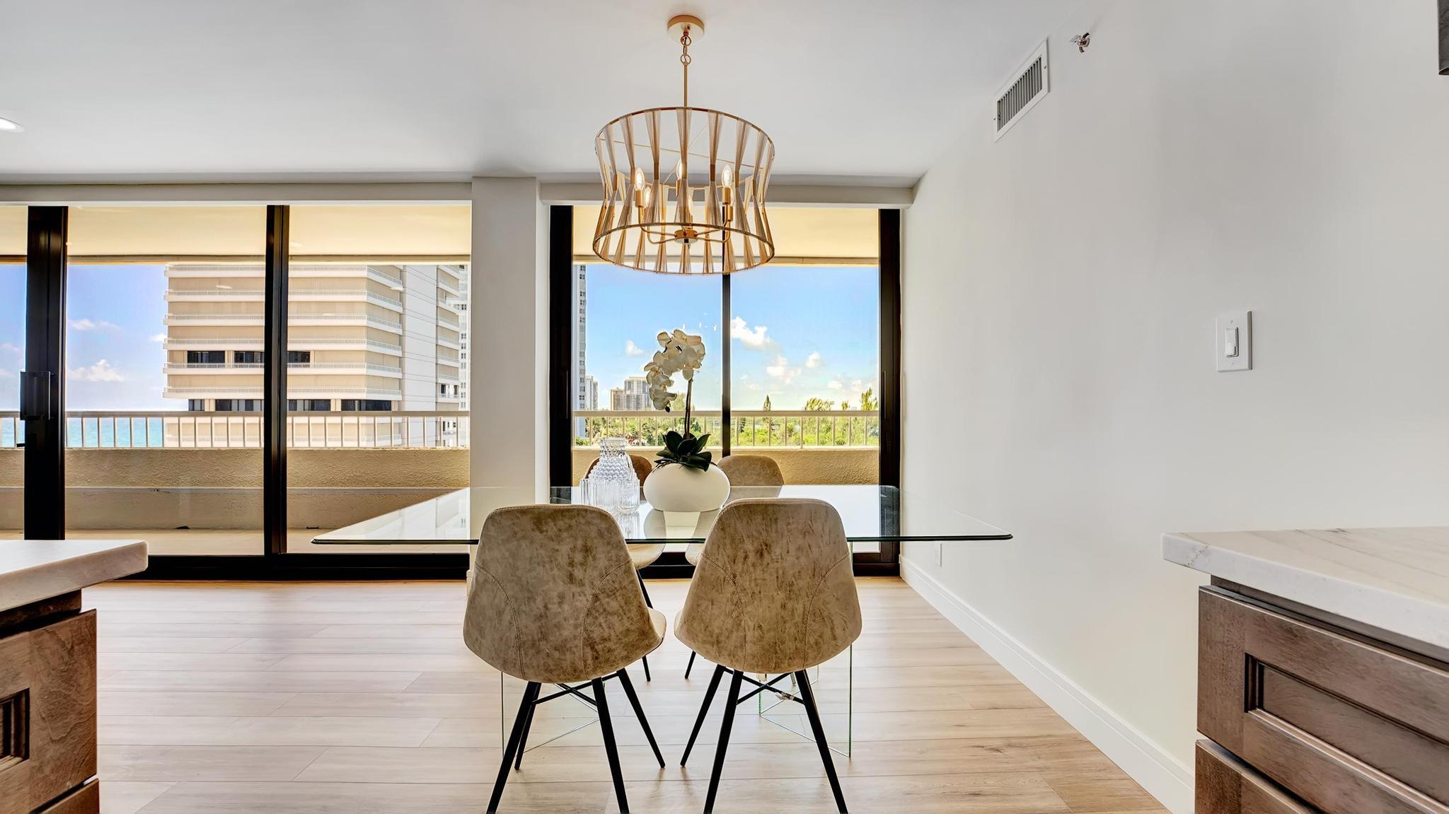 Condo dining area with a glass table, gold chandelier, and balcony overlooking neighboring towers and ocean water.