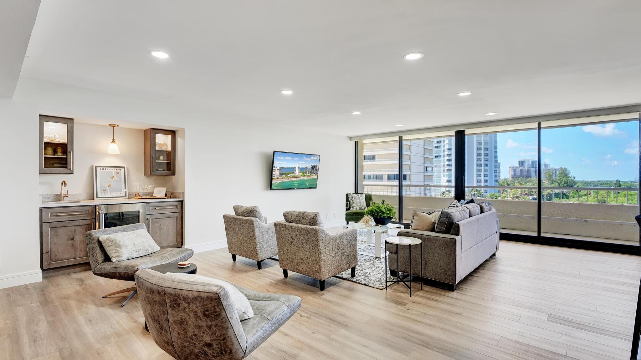 Furnished condo living room with a wet bar and floor-to-ceiling windows overlooking the water and high-rises.