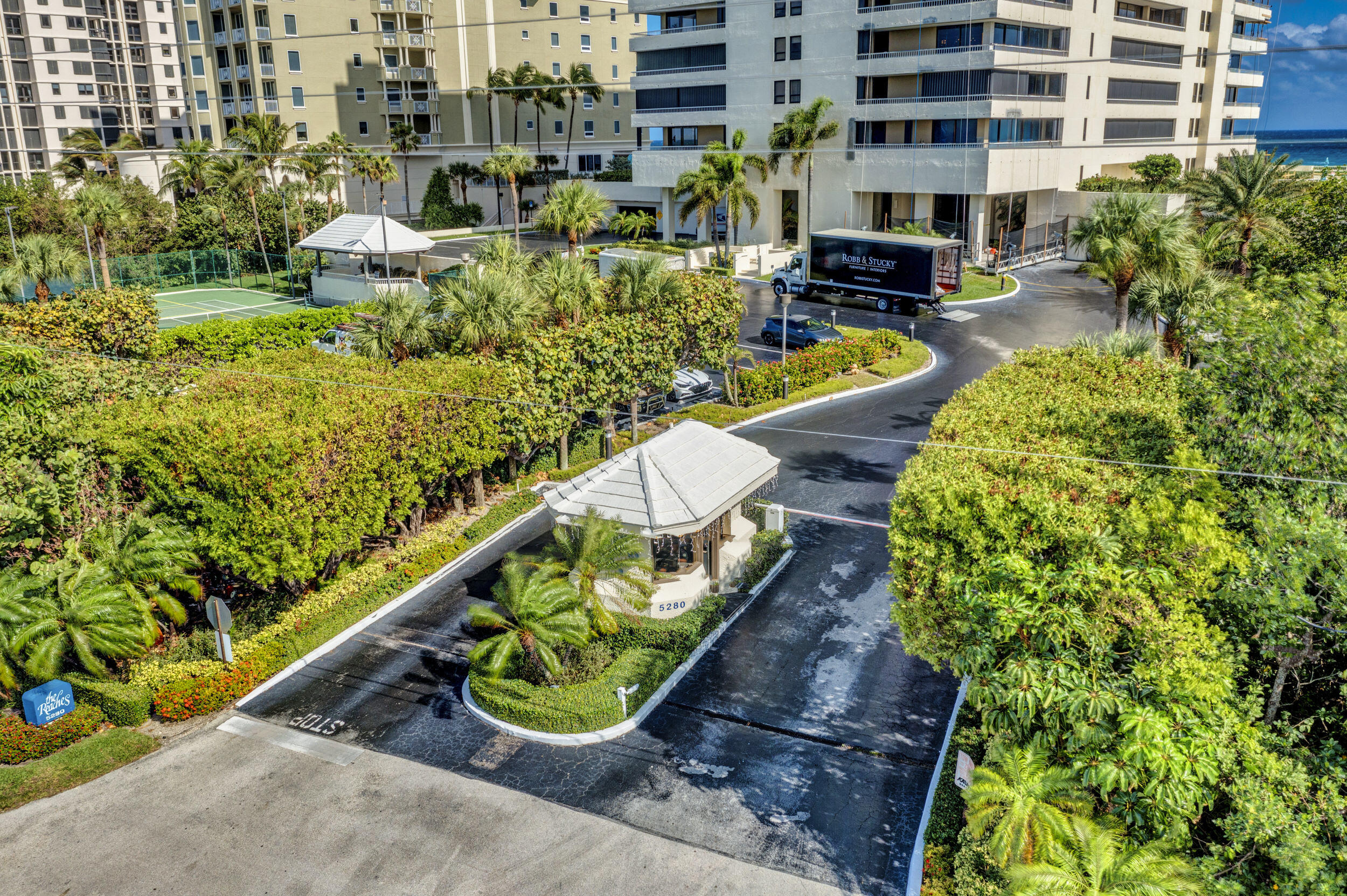 Aerial view of a palm-lined condominium entrance with a tennis court and ocean visible beyond.
