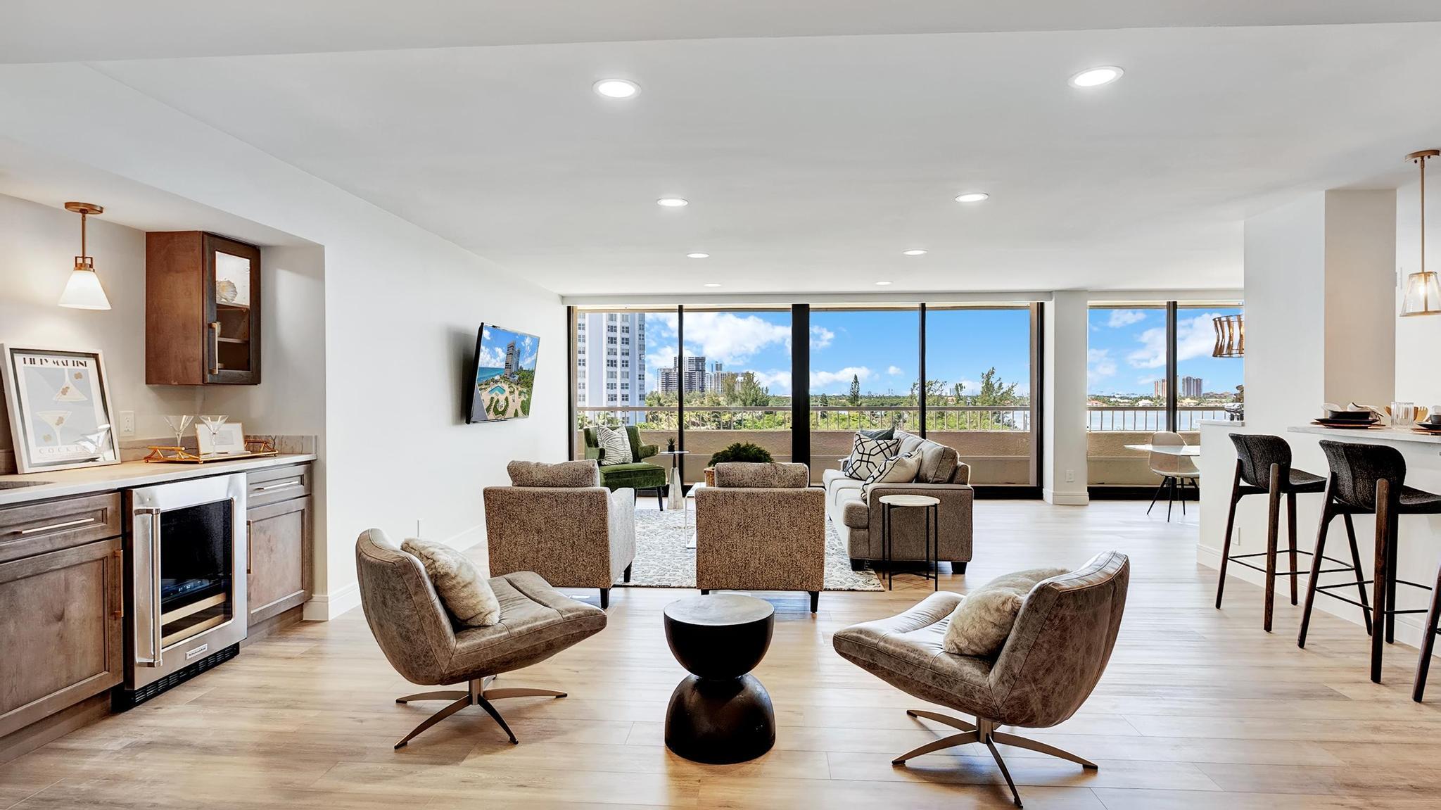 Furnished open-plan living room with floor-to-ceiling windows overlooking water and nearby buildings.