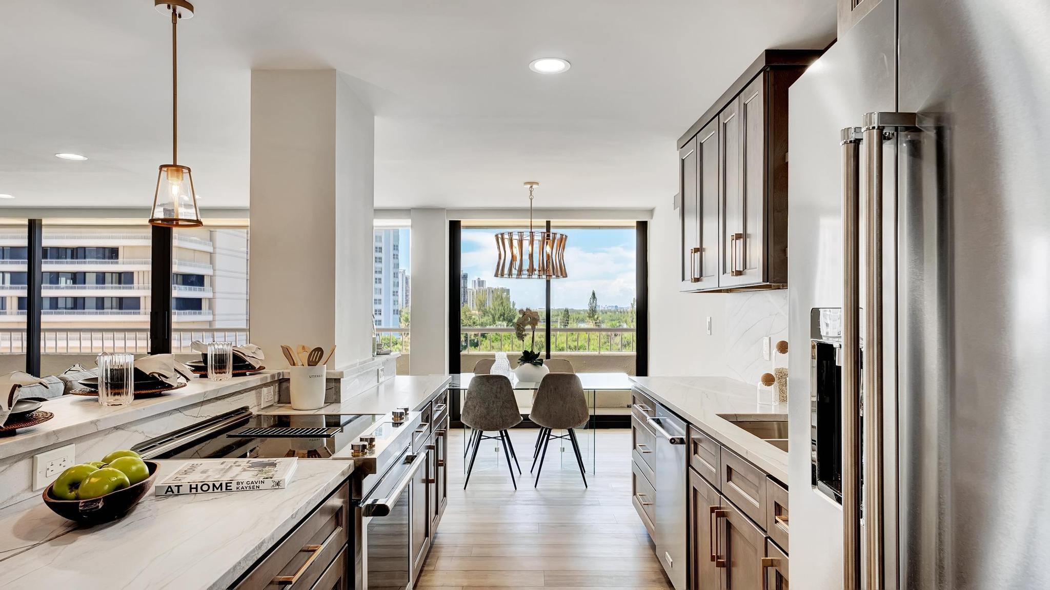 Modern kitchen with marble counters, dark wood cabinets, and windows overlooking greenery and nearby buildings.