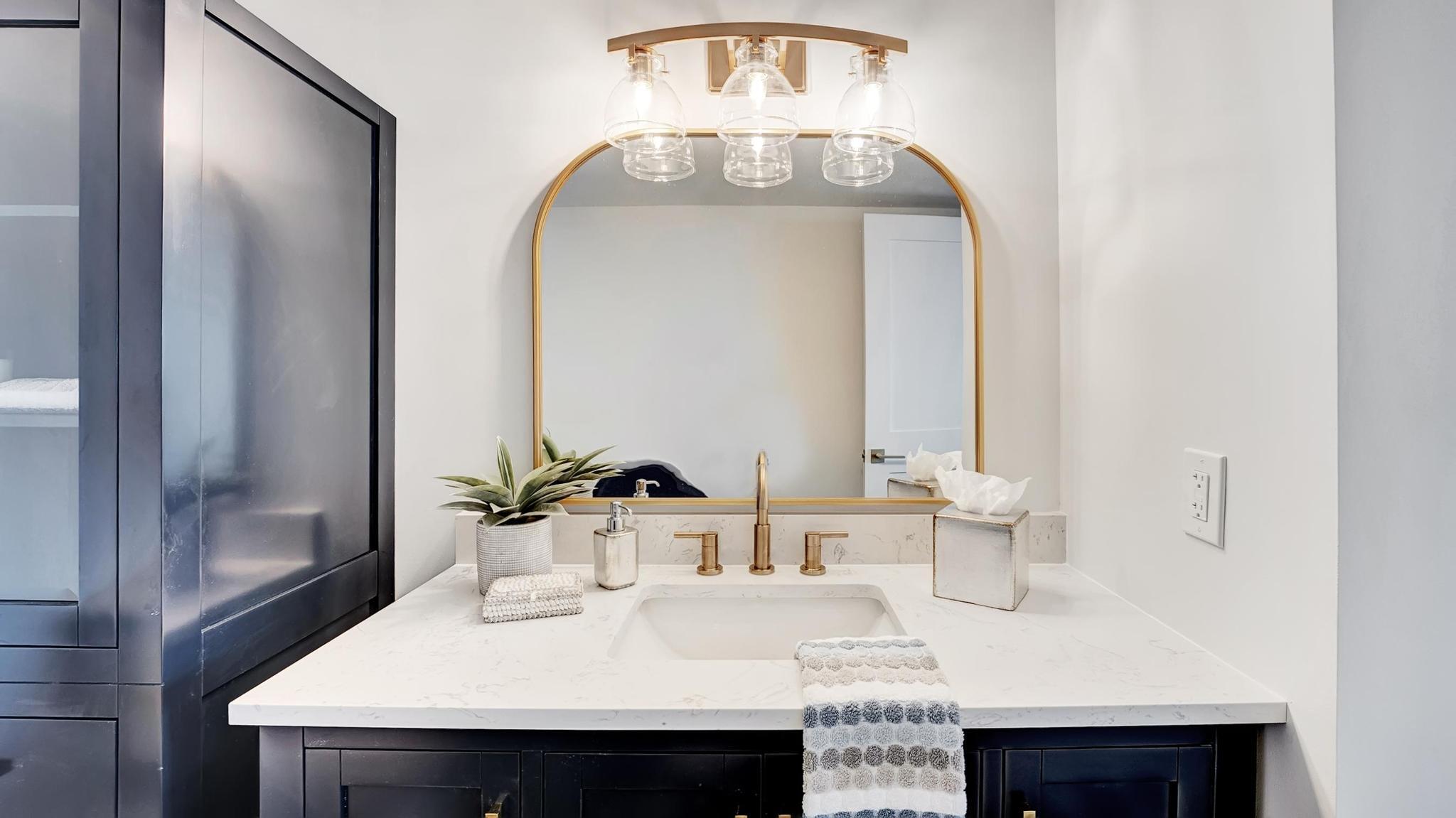 Bathroom vanity with navy cabinetry, white stone counter, gold faucet, and arched mirror.