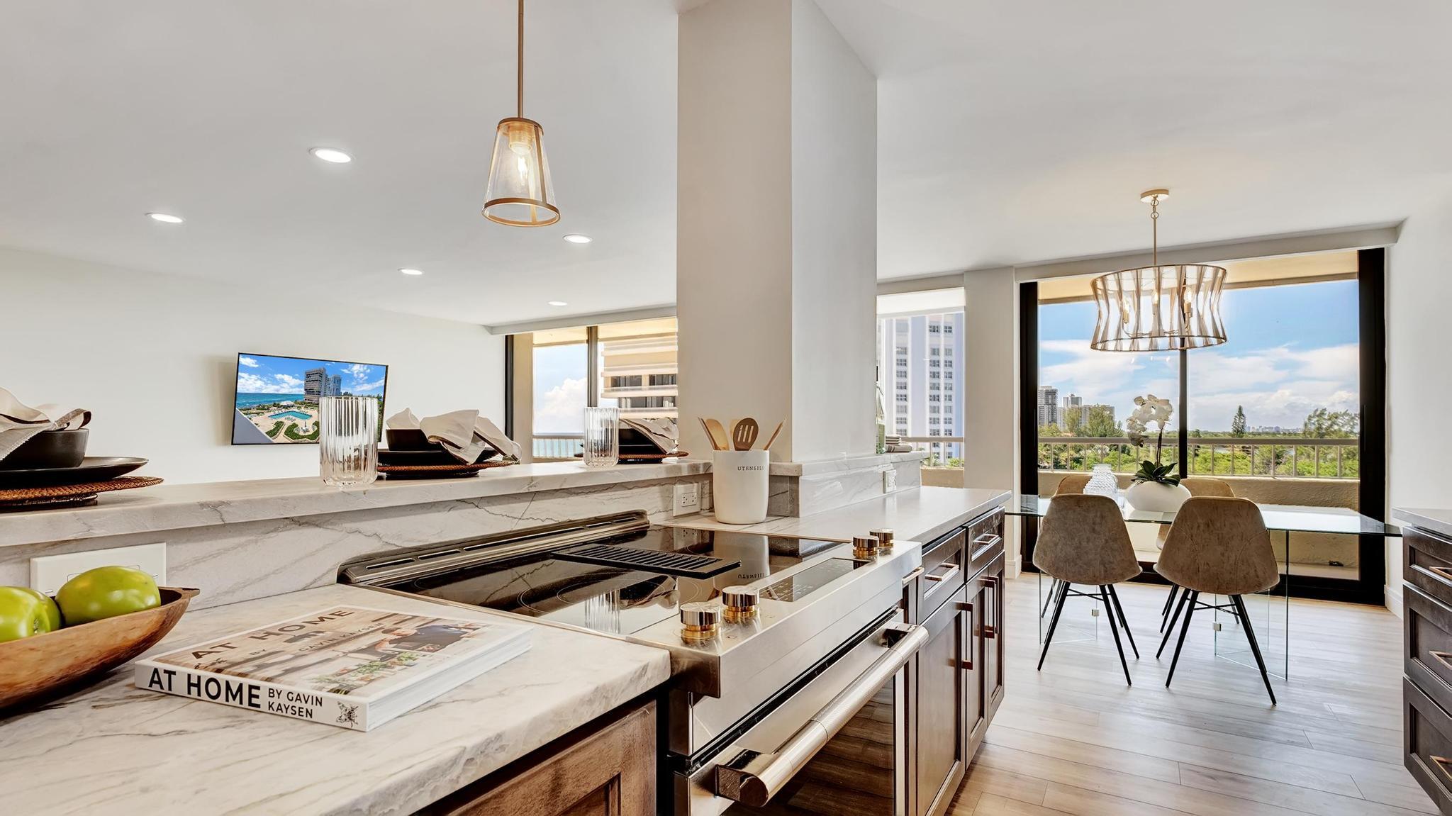 Marble-island kitchen with built-in cooktop and dining table beside floor-to-ceiling balcony windows.