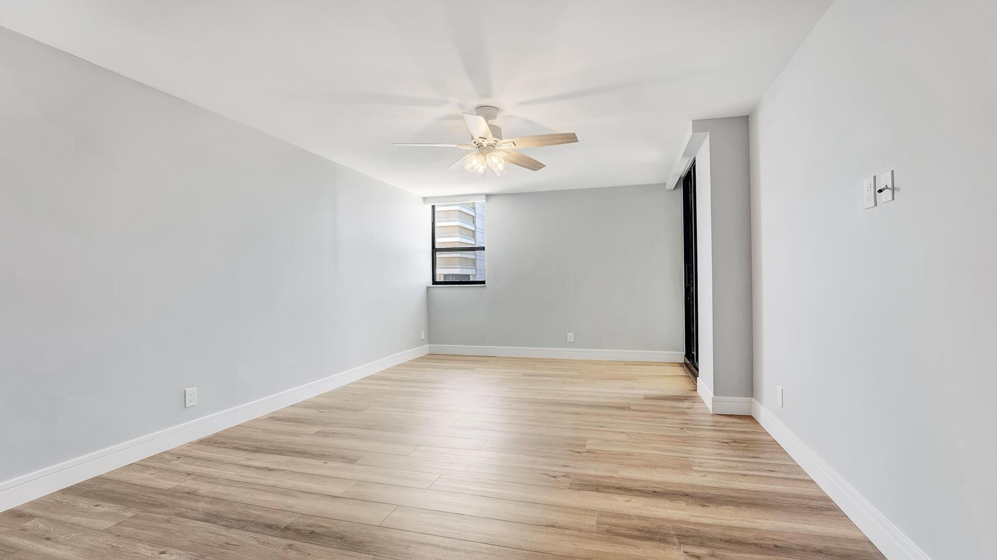 Empty condo room with wood-look tile floors, a ceiling fan, and a window facing a neighboring building.