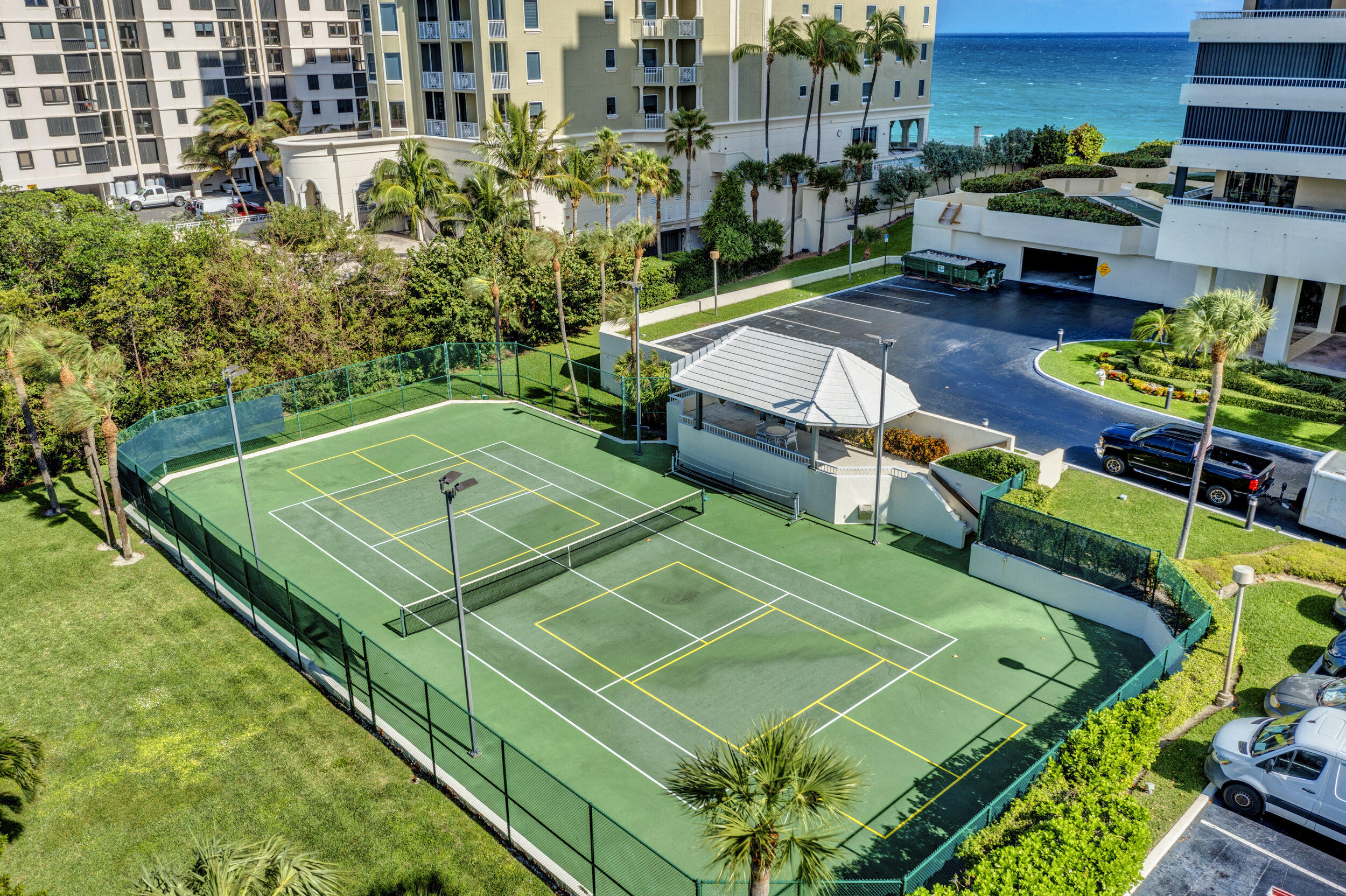 Two fenced tennis courts beside a beachfront condominium, with palm trees and ocean beyond.