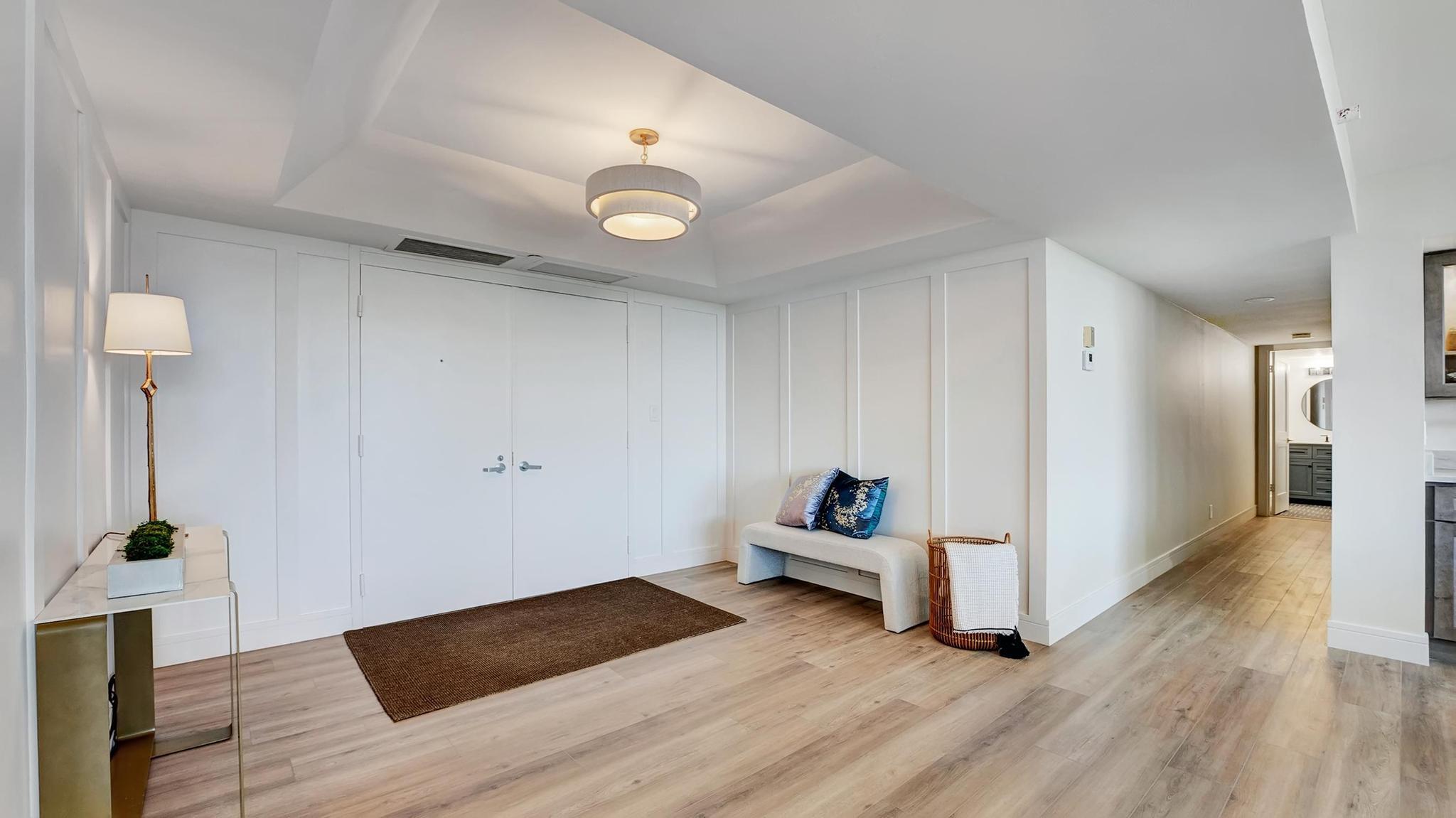 Bright condo entryway with a white bench, blue pillows, and a hallway leading to a bathroom.