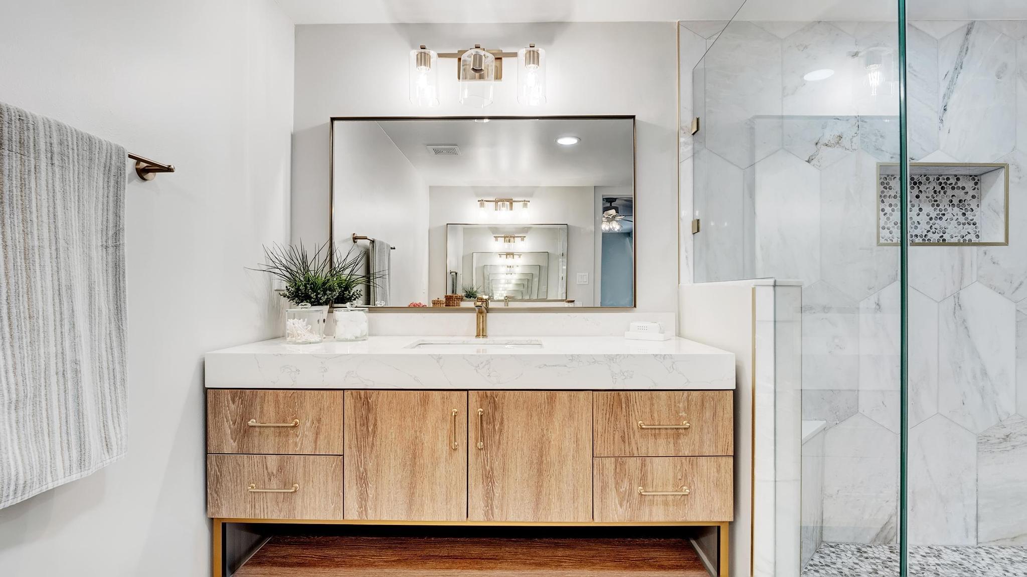 Modern bathroom with a wood-grain vanity, brass fixtures, and glass-enclosed marble shower.