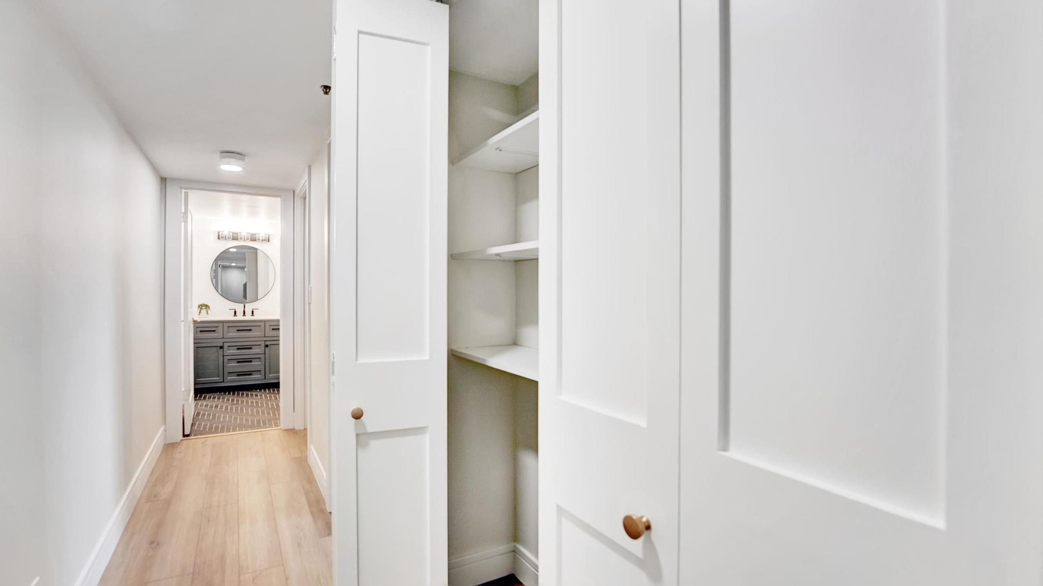 Apartment hallway with open white closet shelving and a vanity with a round mirror at the end.