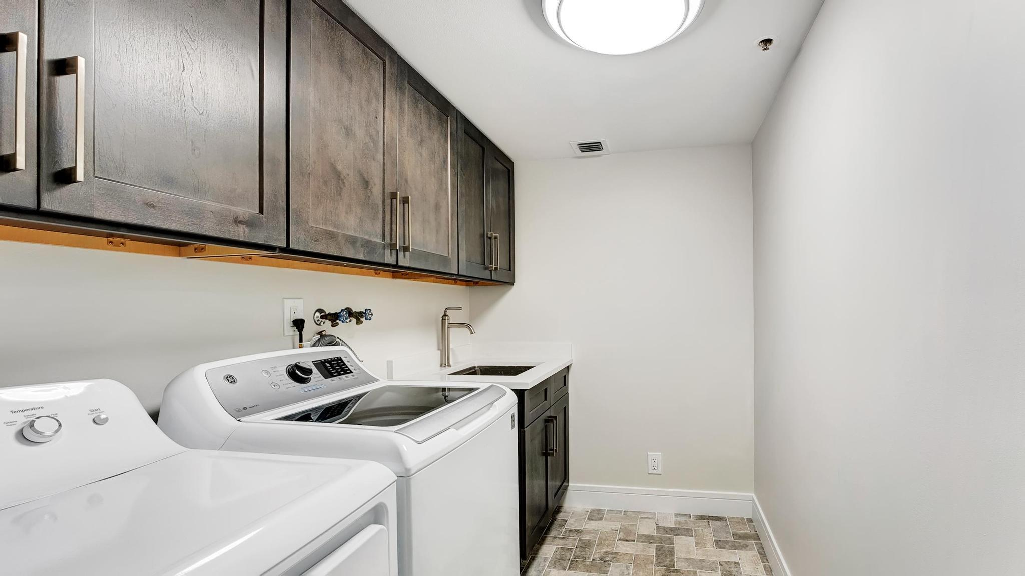 Laundry room with white washer and dryer, dark cabinets, utility sink, and patterned tile floor.