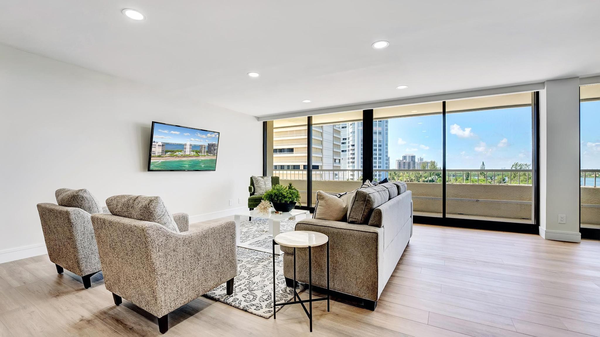 Furnished living room with floor-to-ceiling windows overlooking water and neighboring high-rises.