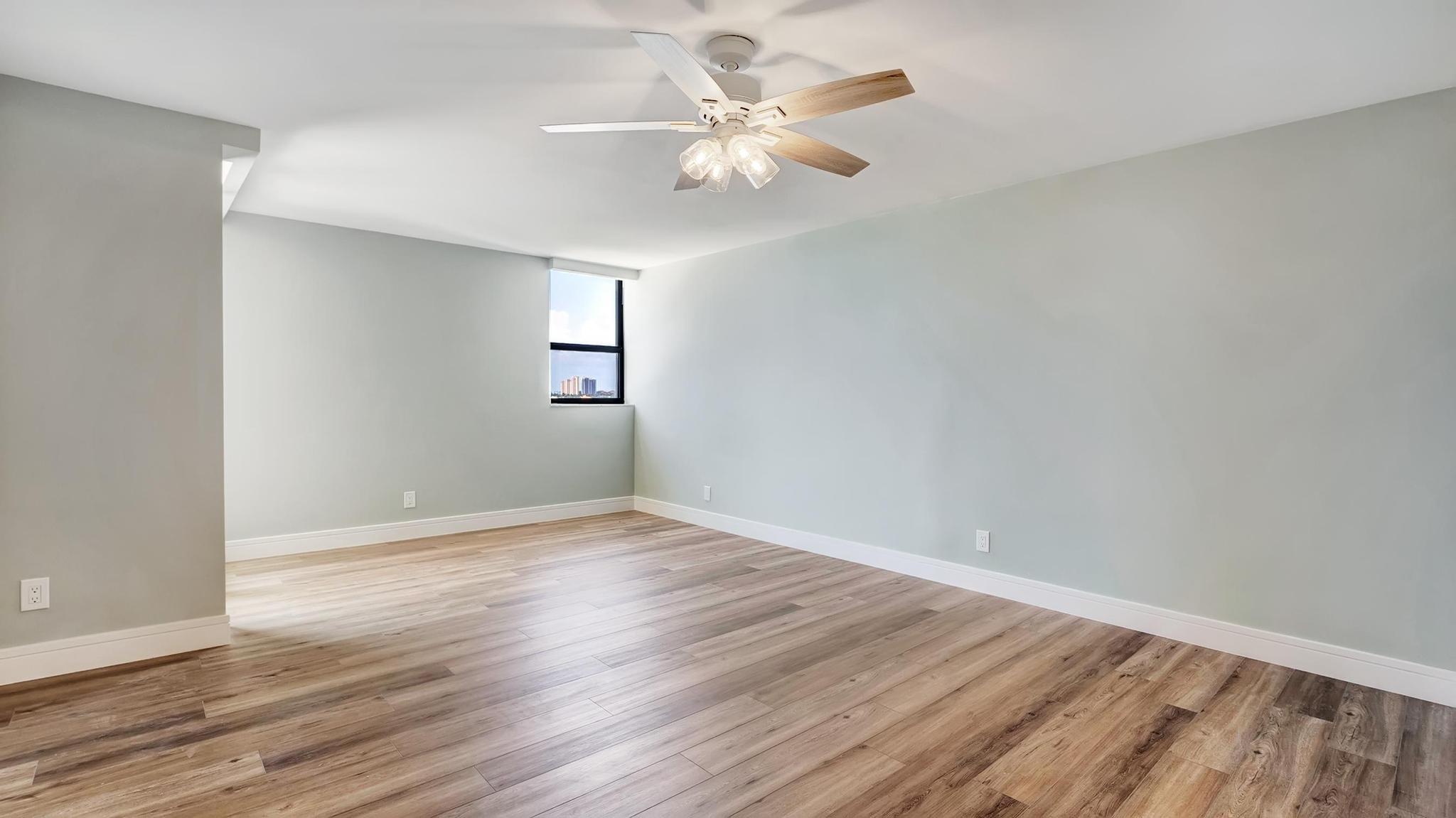 Vacant room with wood-look floors, pale gray walls, ceiling fan, and a small window with distant buildings.