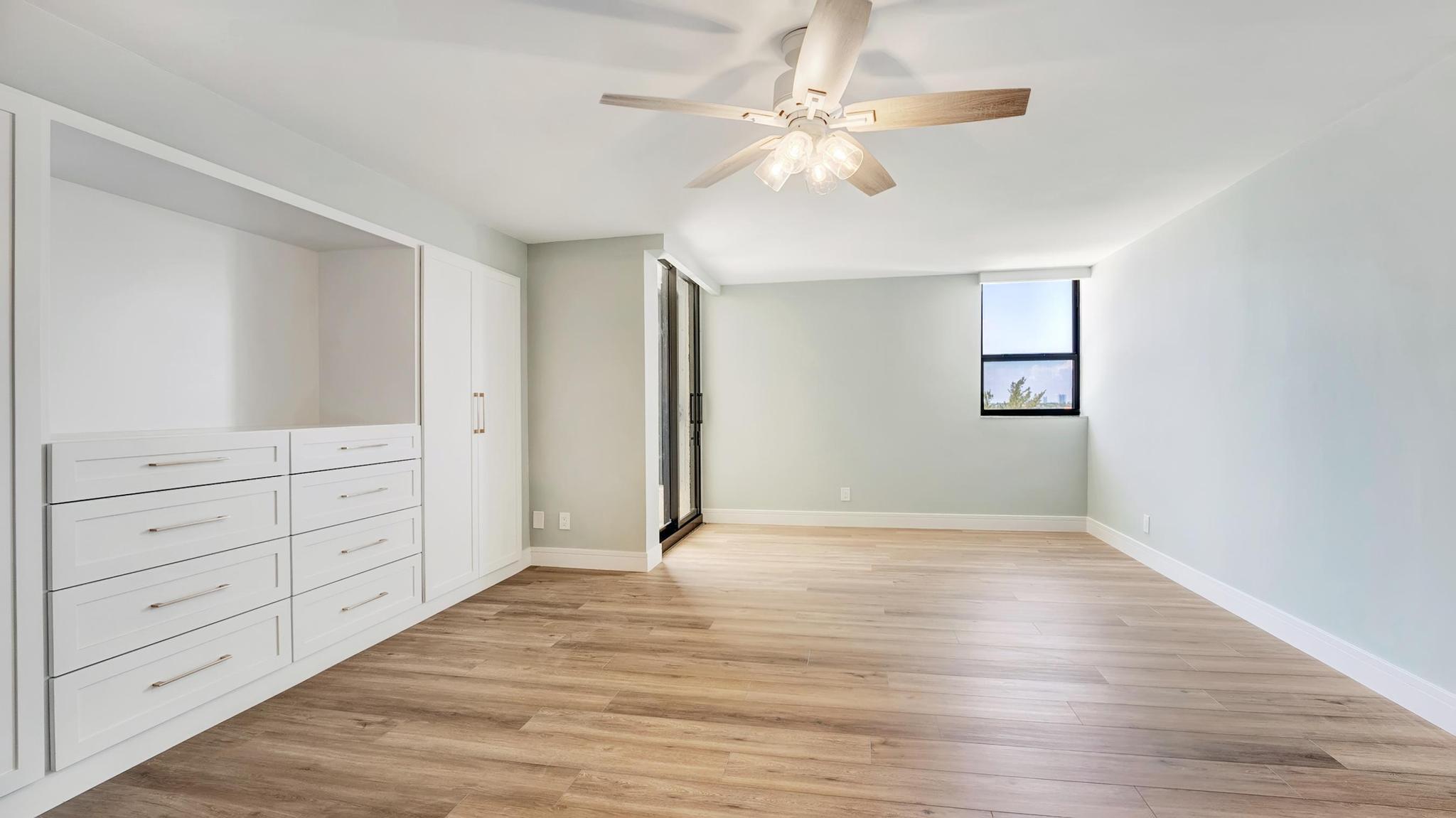 Empty bedroom with built-in white drawers, wood-look flooring, and a ceiling fan.