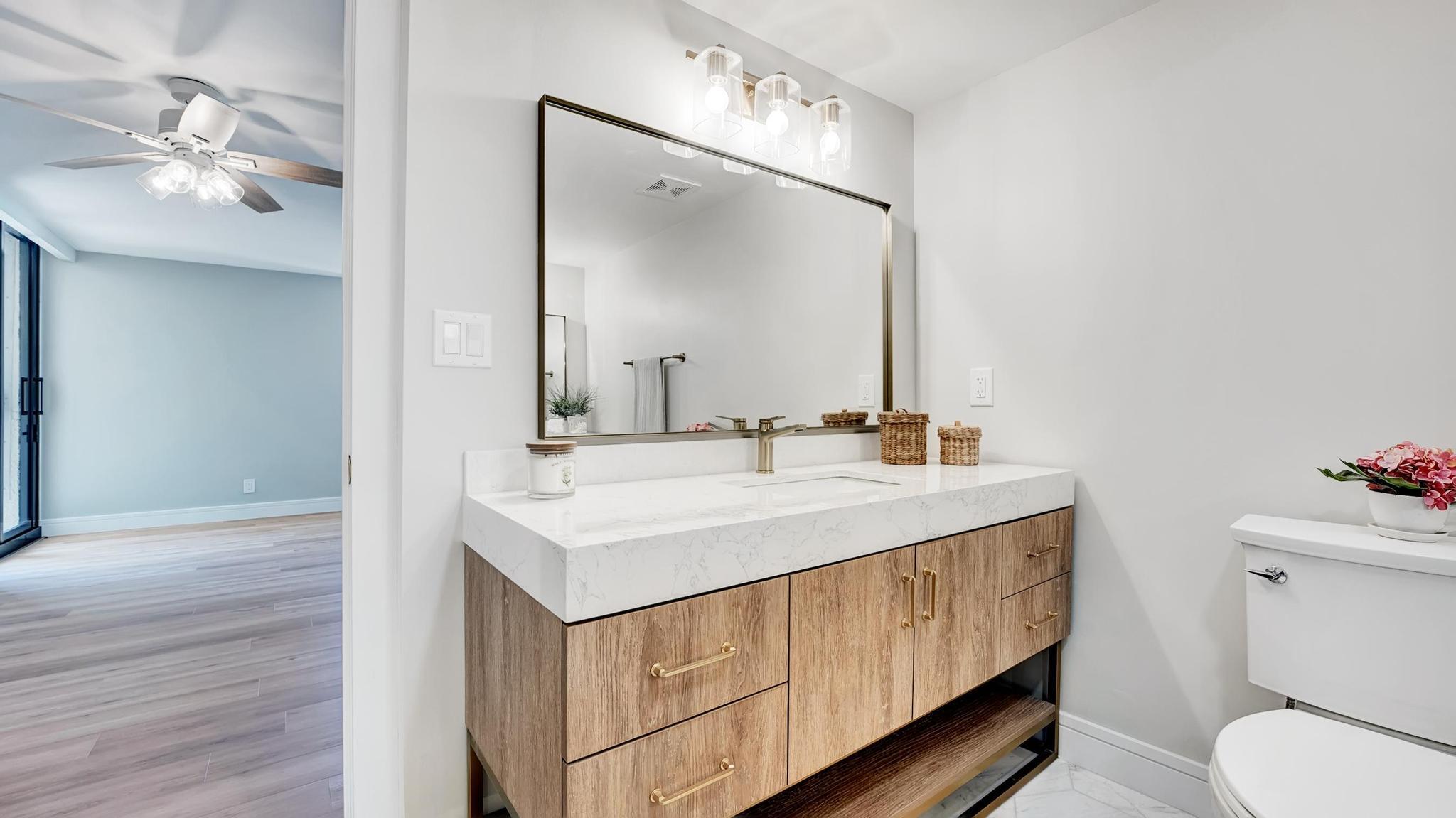 Bathroom with a wood vanity, white countertop, brass fixtures, and a large mirror beside an open doorway.