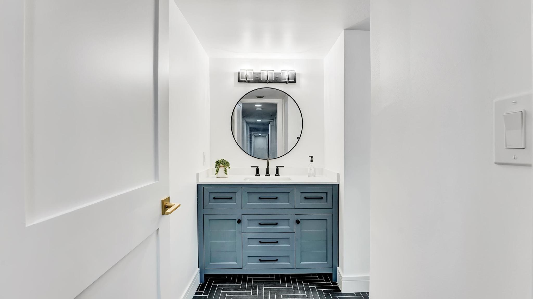 Bathroom vanity with blue-gray cabinetry, a round mirror, and dark herringbone floor tile.