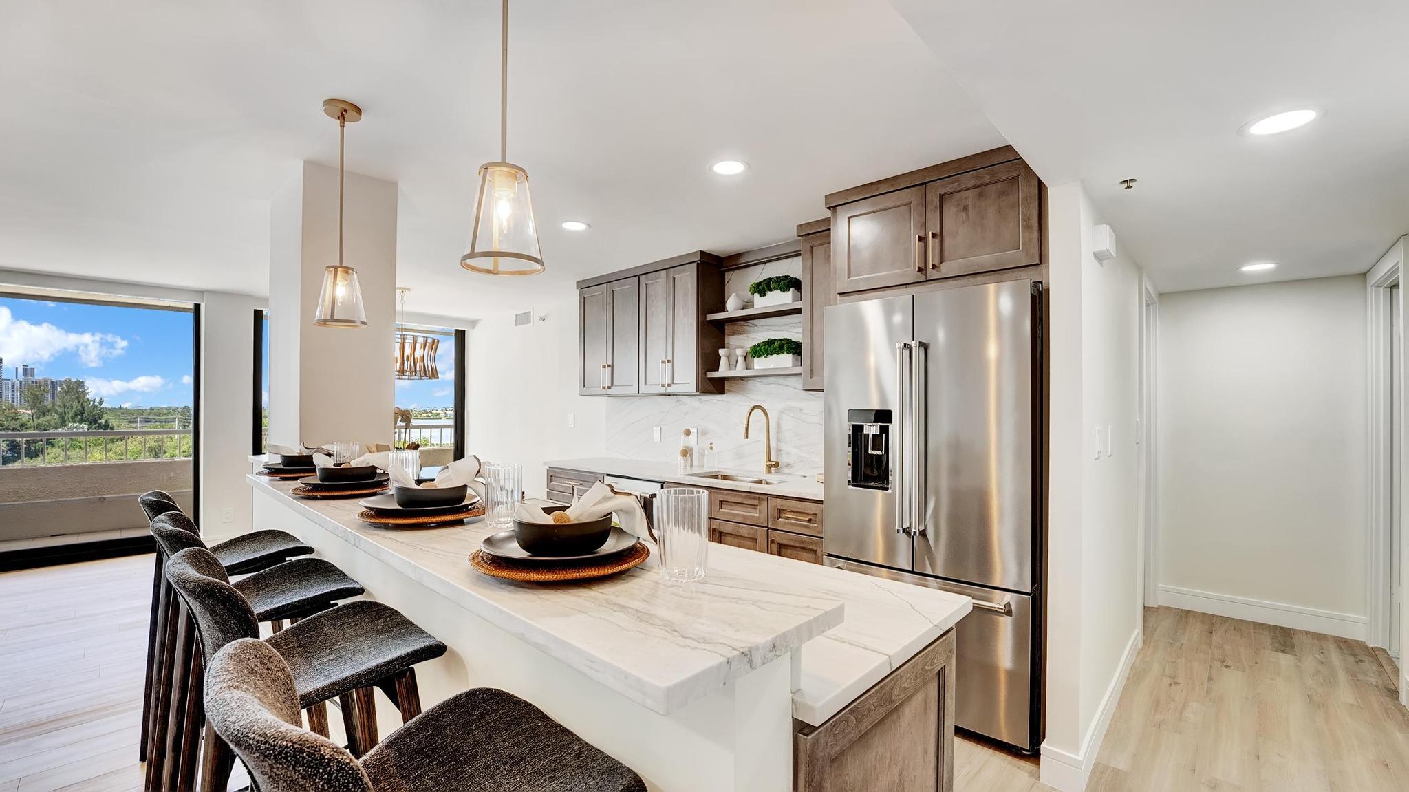 Staged condo kitchen with marble island, dark bar stools, stainless appliances, and balcony views.