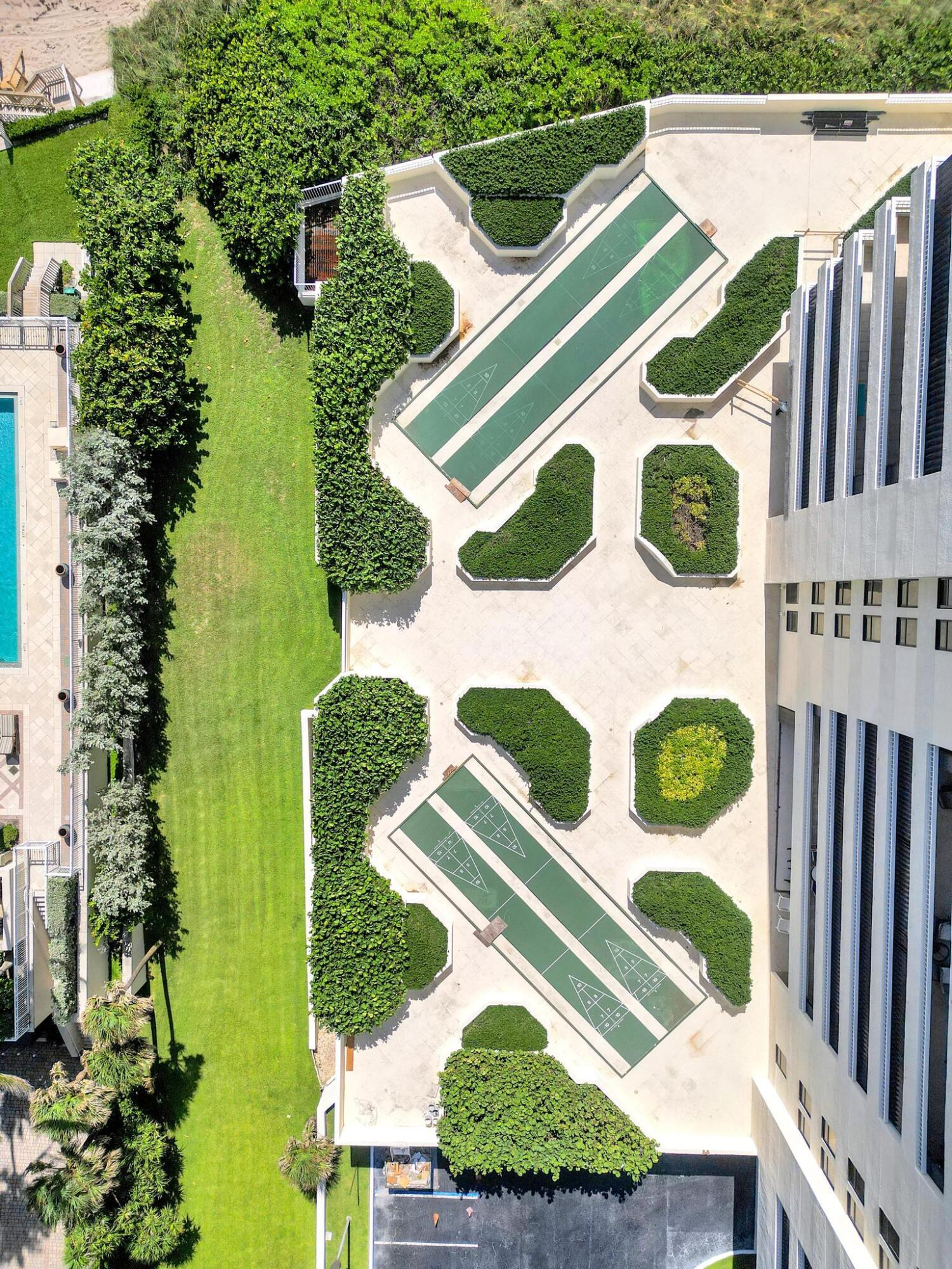 Aerial view of a condominium courtyard with shuffleboard courts, landscaped hedges, a lawn, and a pool.