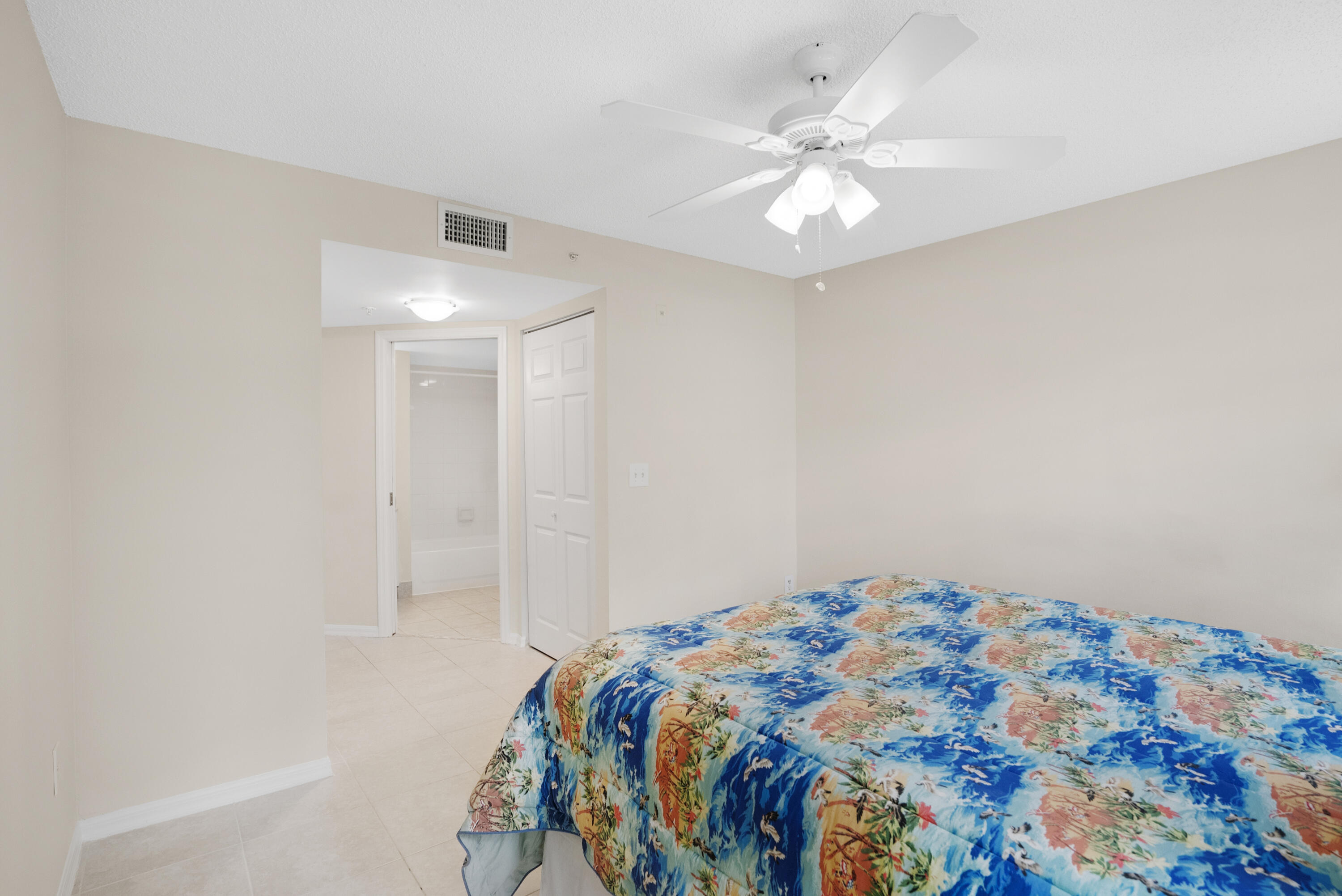 Bedroom with a colorful tropical-print bedspread, ceiling fan, and open doorway to a bathroom.