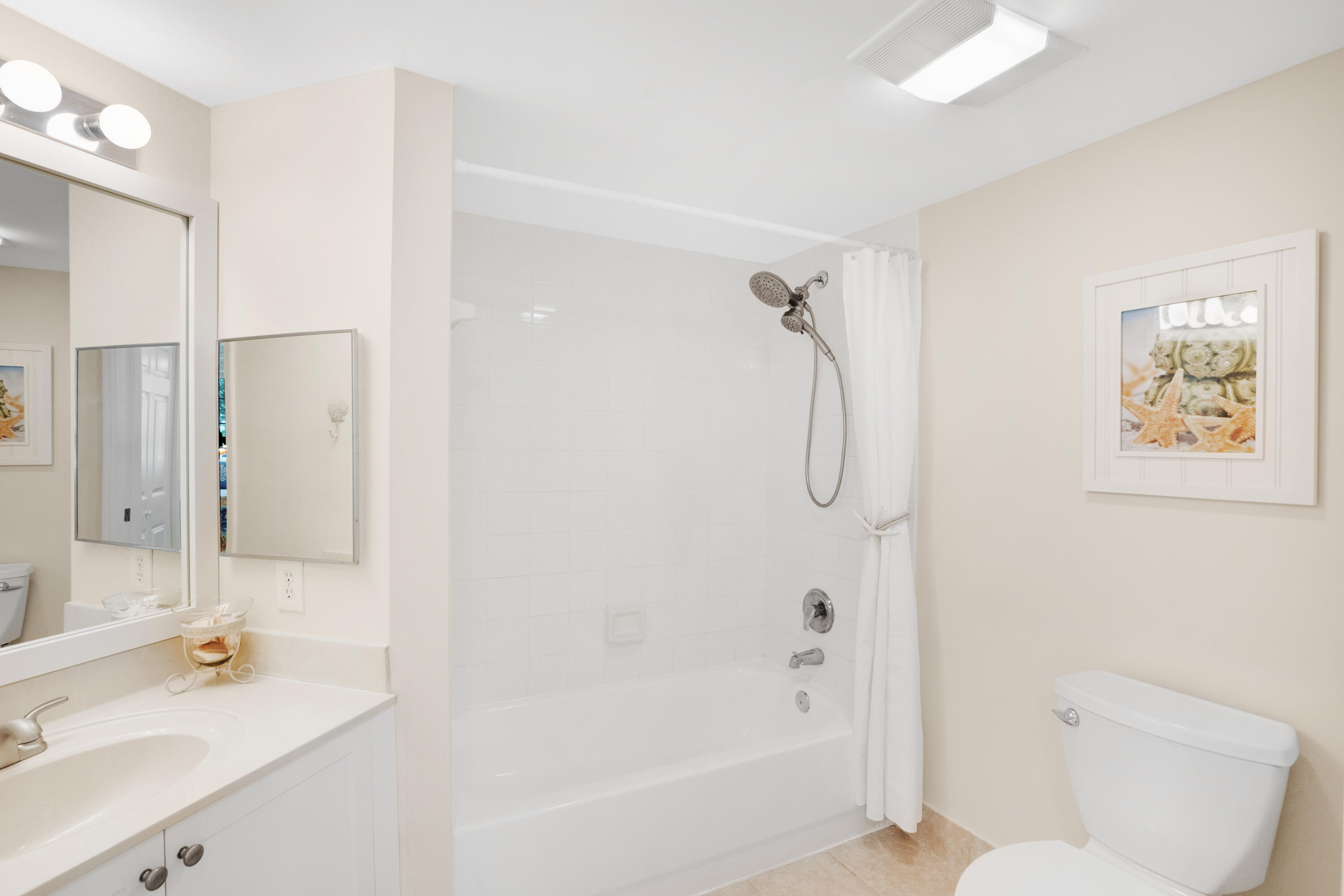 Bathroom with a white vanity, tub and shower, toilet, beige tile floor, and framed beach artwork.