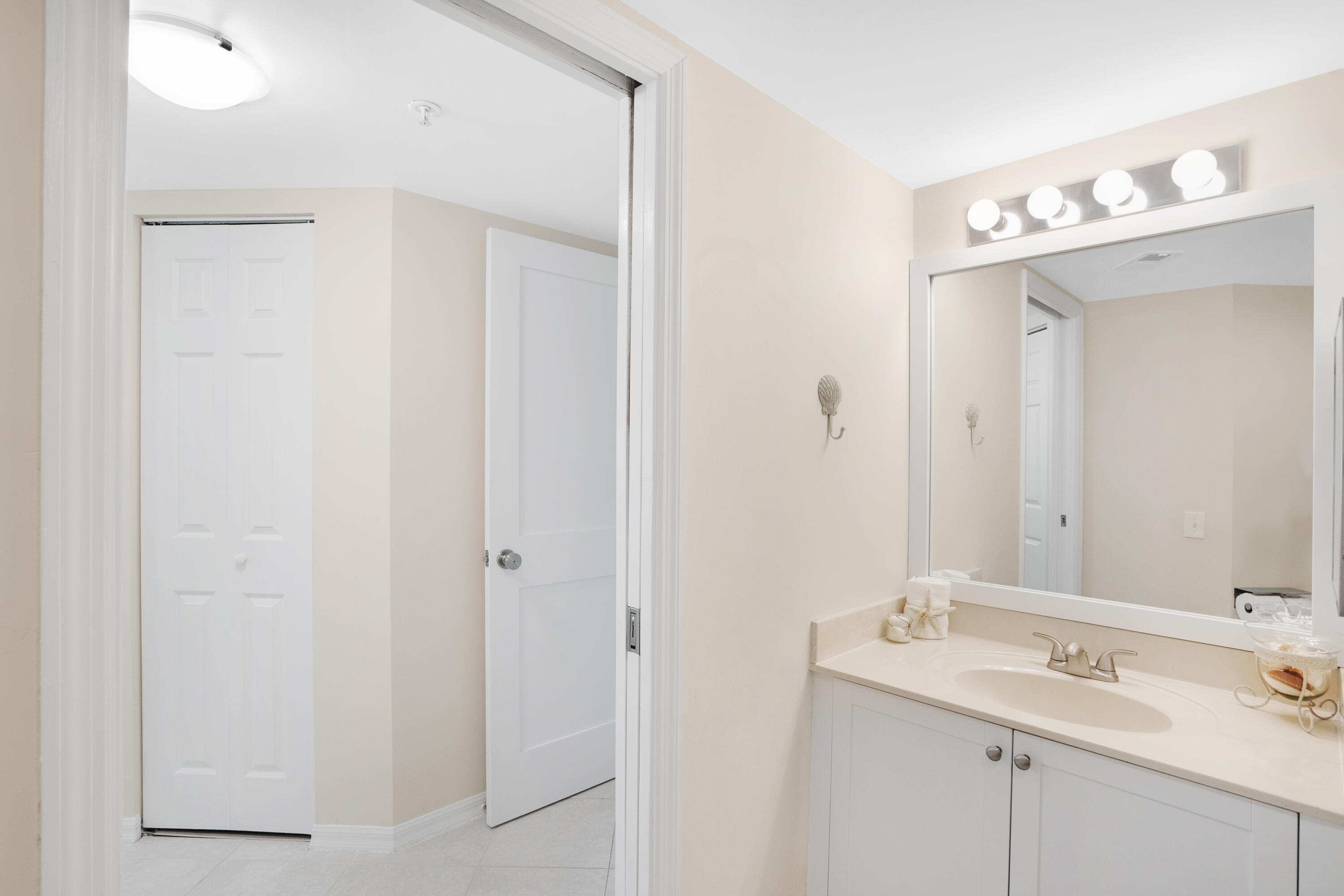 Bathroom vanity with a large mirror and bright lights beside an open doorway to a tiled hall.