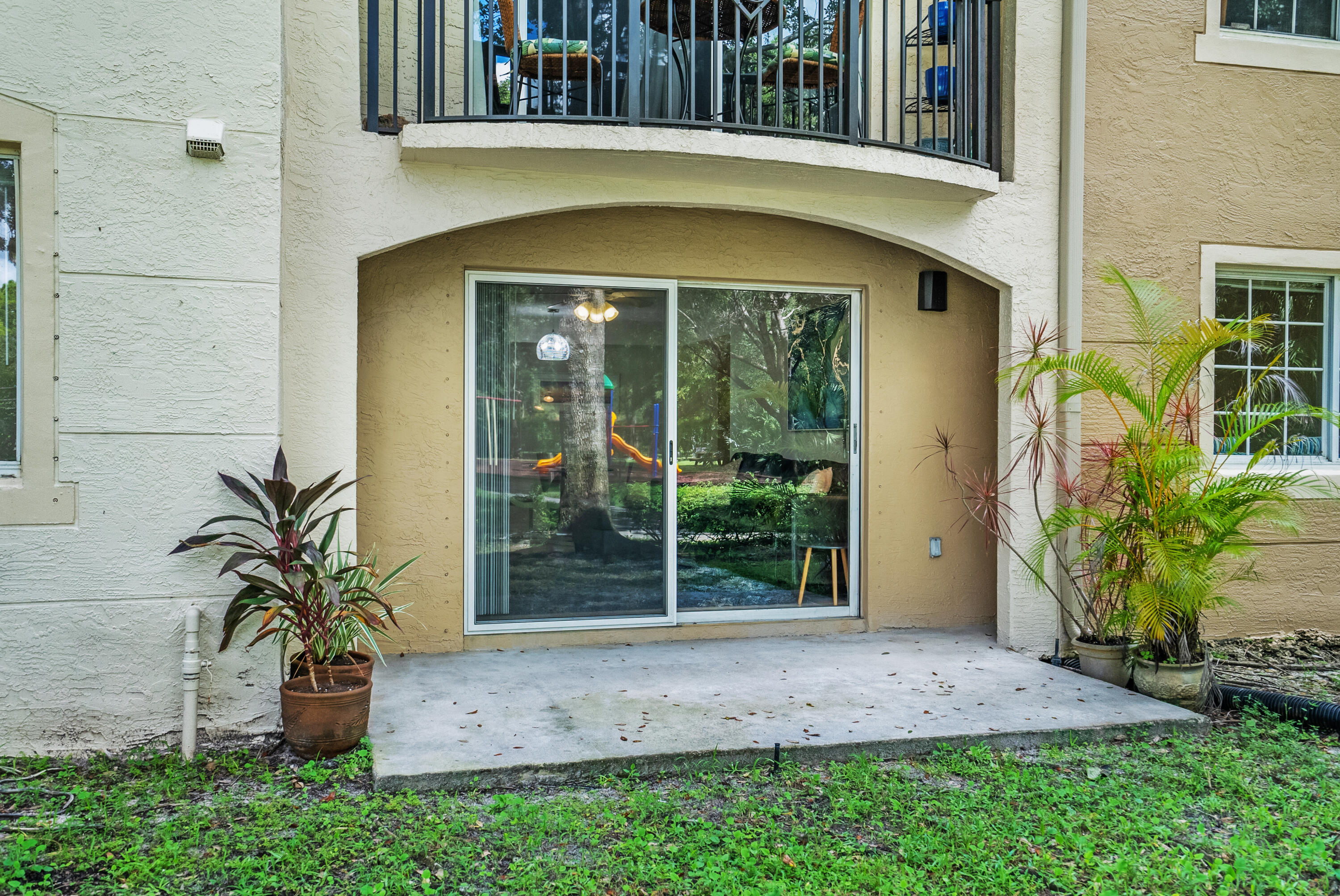 Ground-floor condo patio with sliding glass doors, tropical plants, and a balcony above.