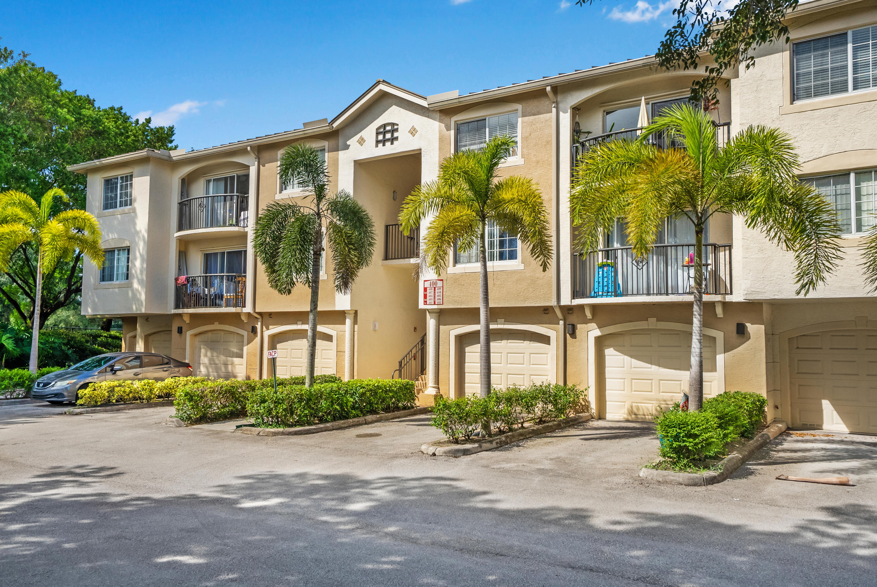 Beige condominium building with balconies, garage doors, and palm trees along the parking area.