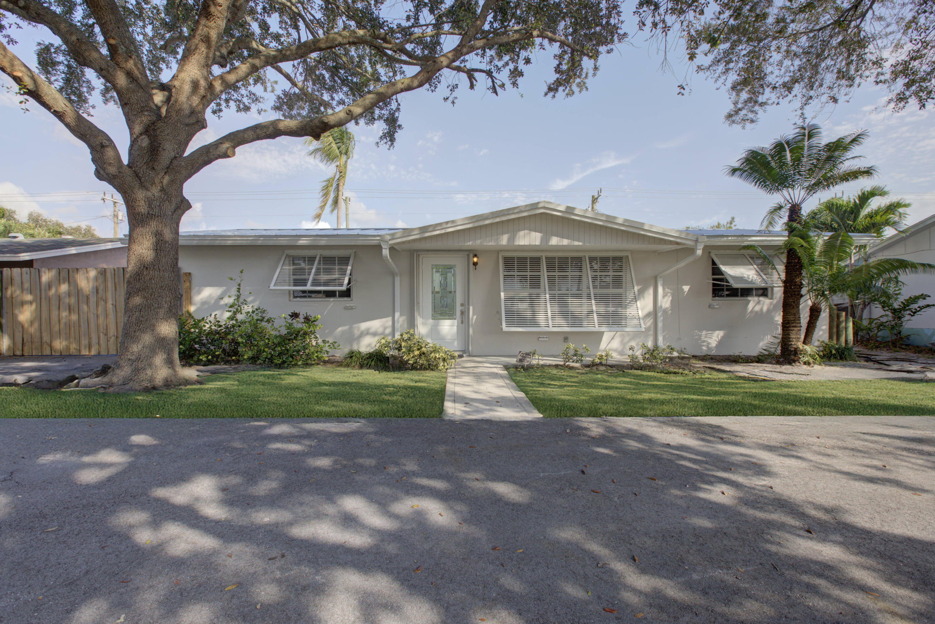 Single-story white house with shuttered windows, a front walkway, large shade tree, and palm trees.