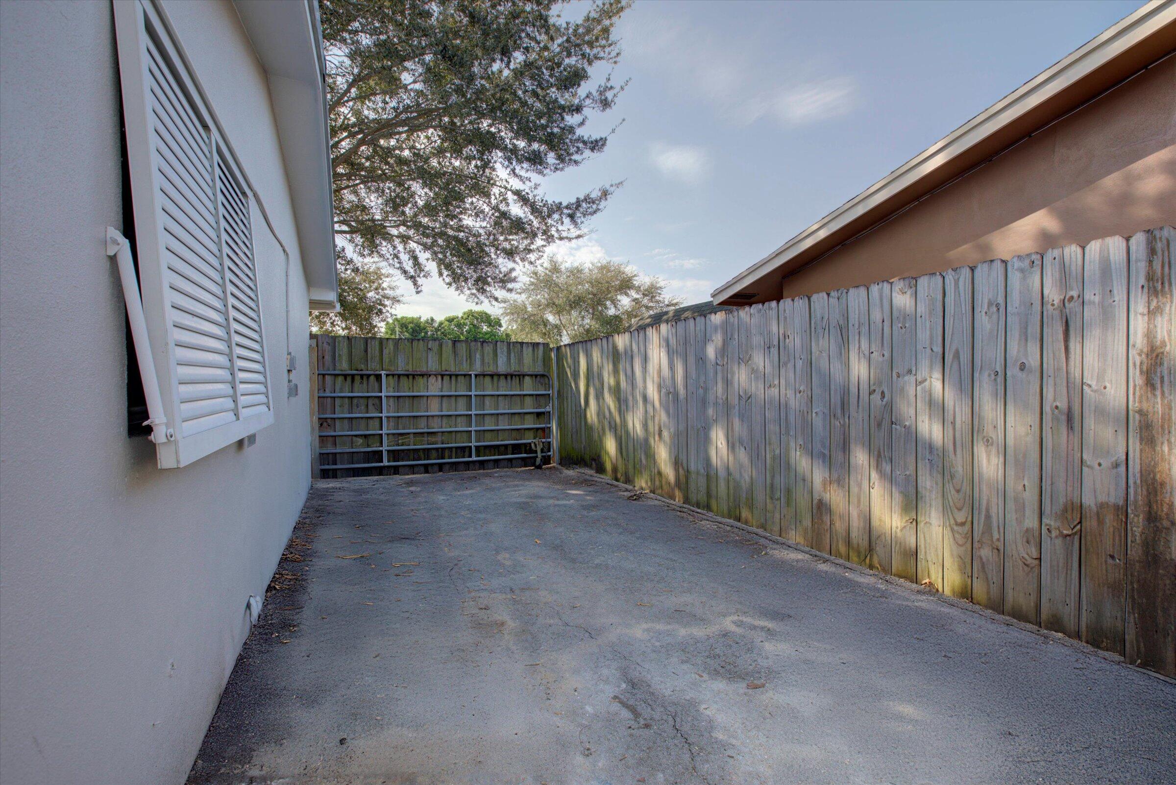 Paved side driveway beside a house, enclosed by wooden fencing and ending at a metal gate.