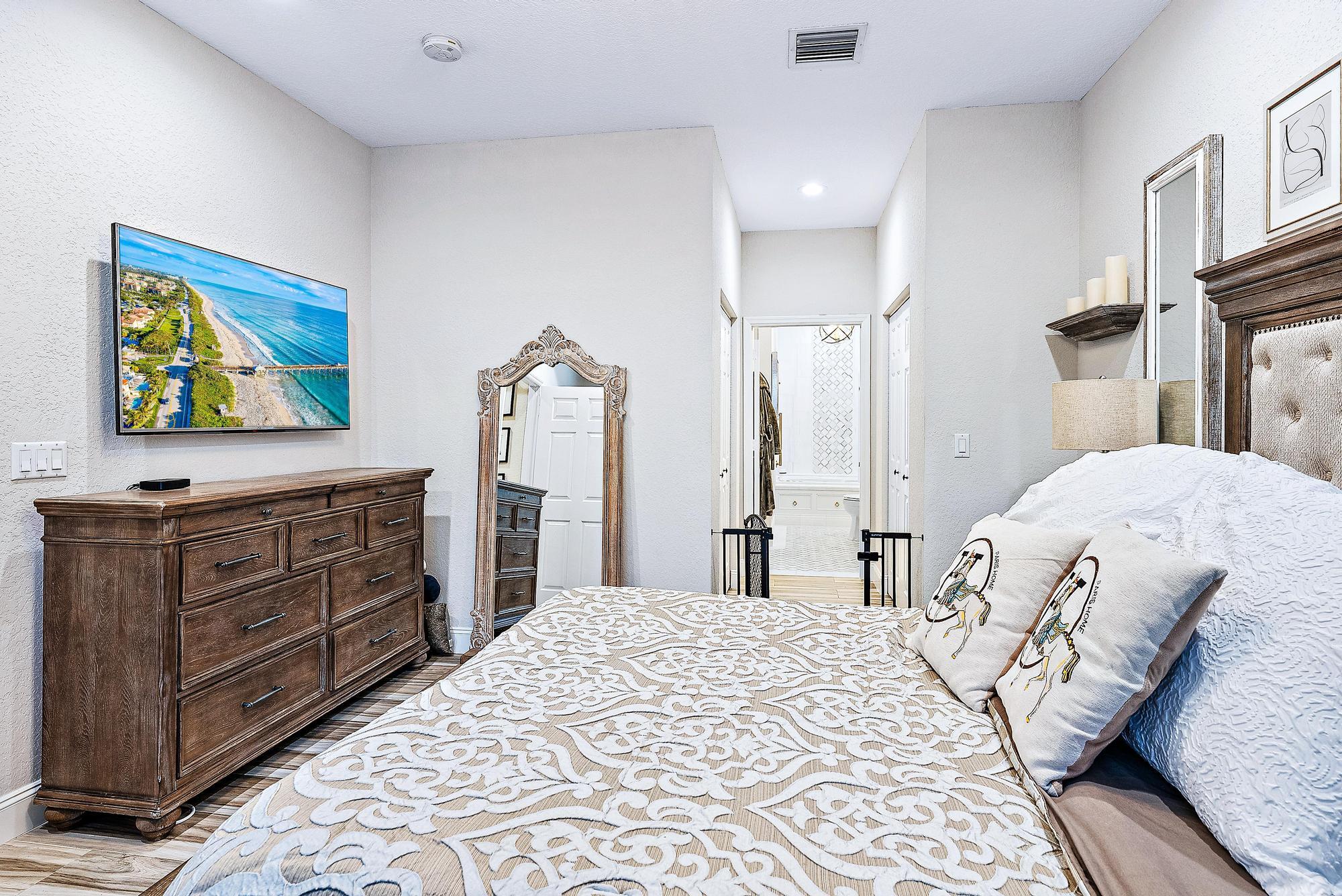 Furnished bedroom with patterned white bedding, dark wood dresser, ornate mirror, and hallway to a bathroom.