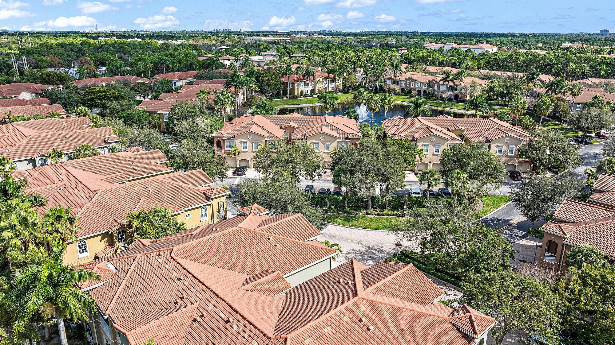 Aerial view of terracotta-roofed homes surrounding a lake and fountain in a palm-lined community.