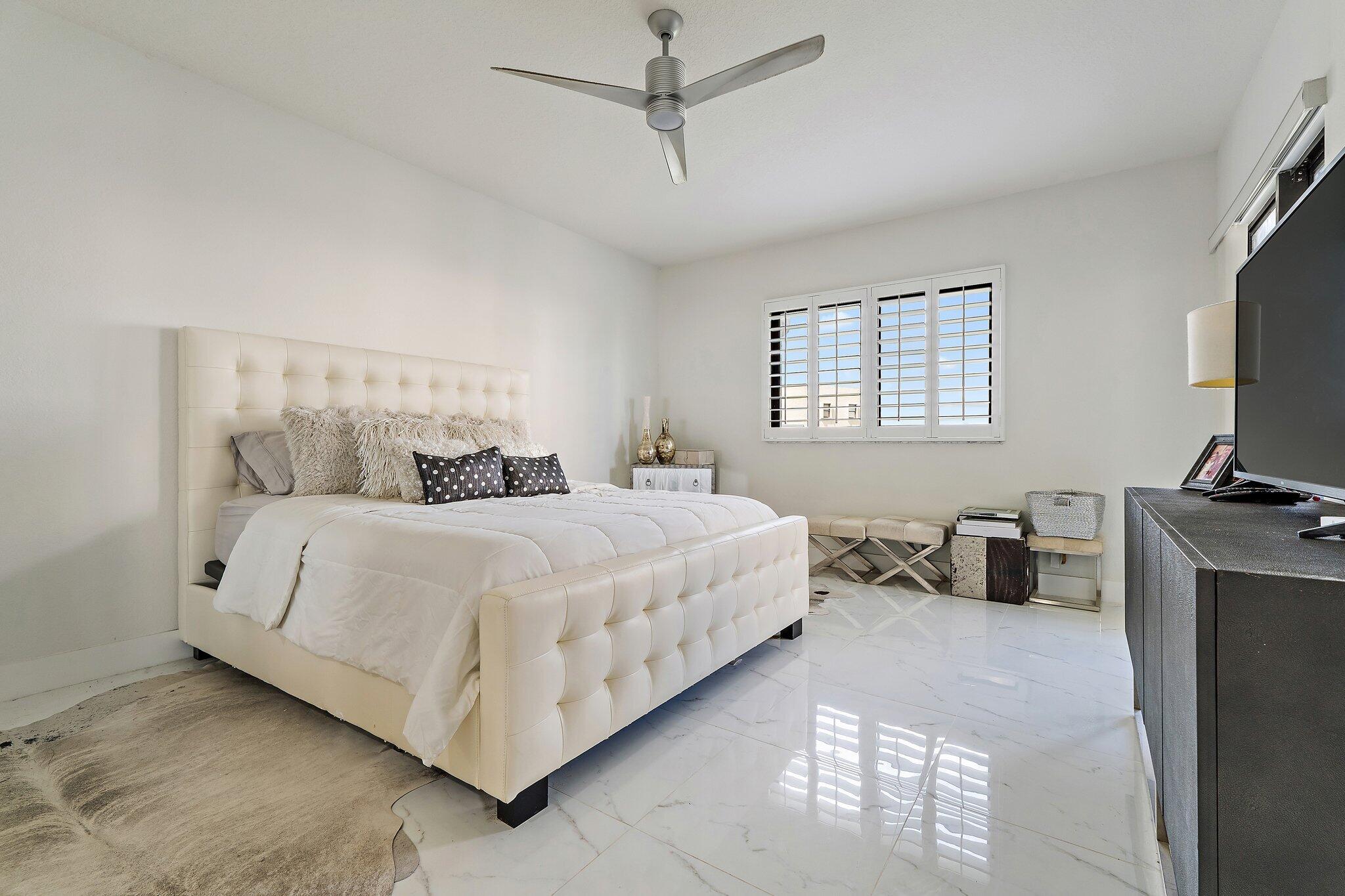 Bedroom with a cream tufted bed, glossy tile floor, and plantation shutters.