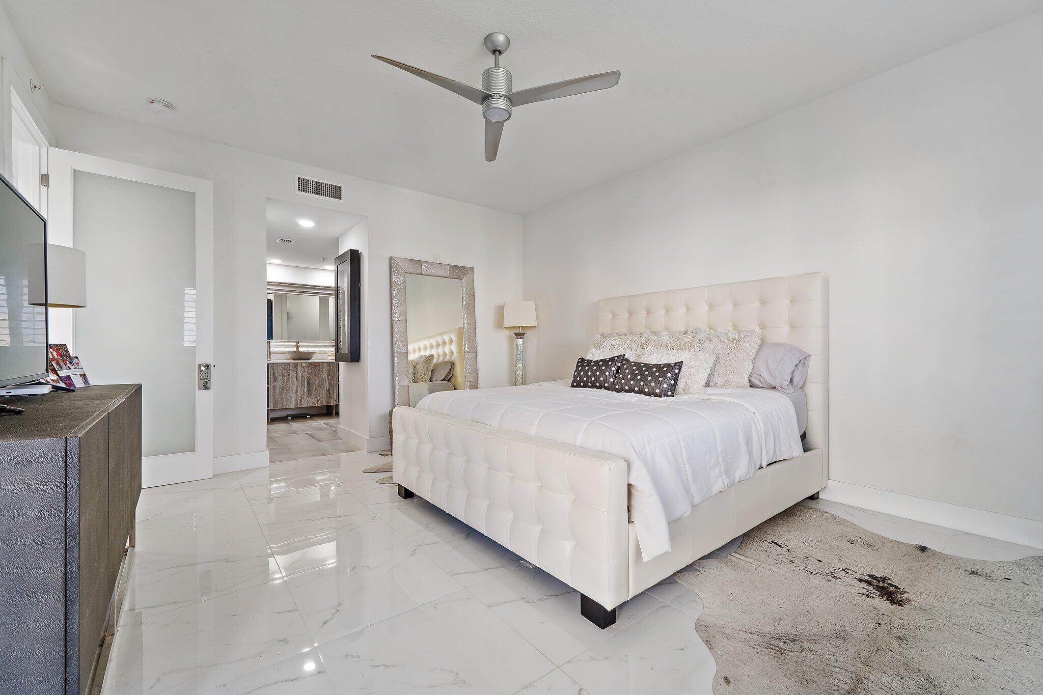 Furnished bedroom with a white tufted bed, ceiling fan, and doorway to an en-suite bathroom.