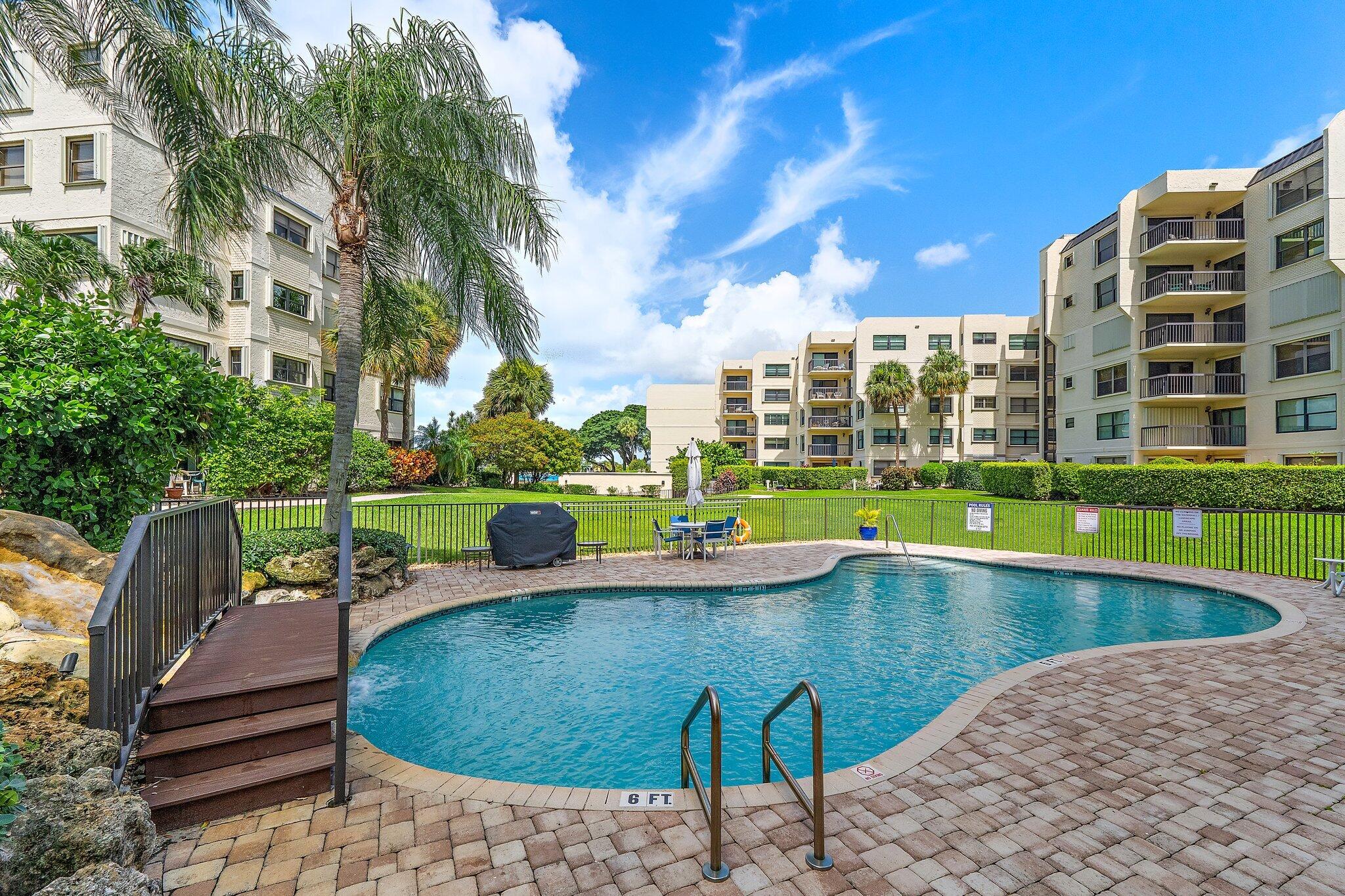 Courtyard pool with a curved deck, wooden access ramp, and palm-lined condominium buildings.
