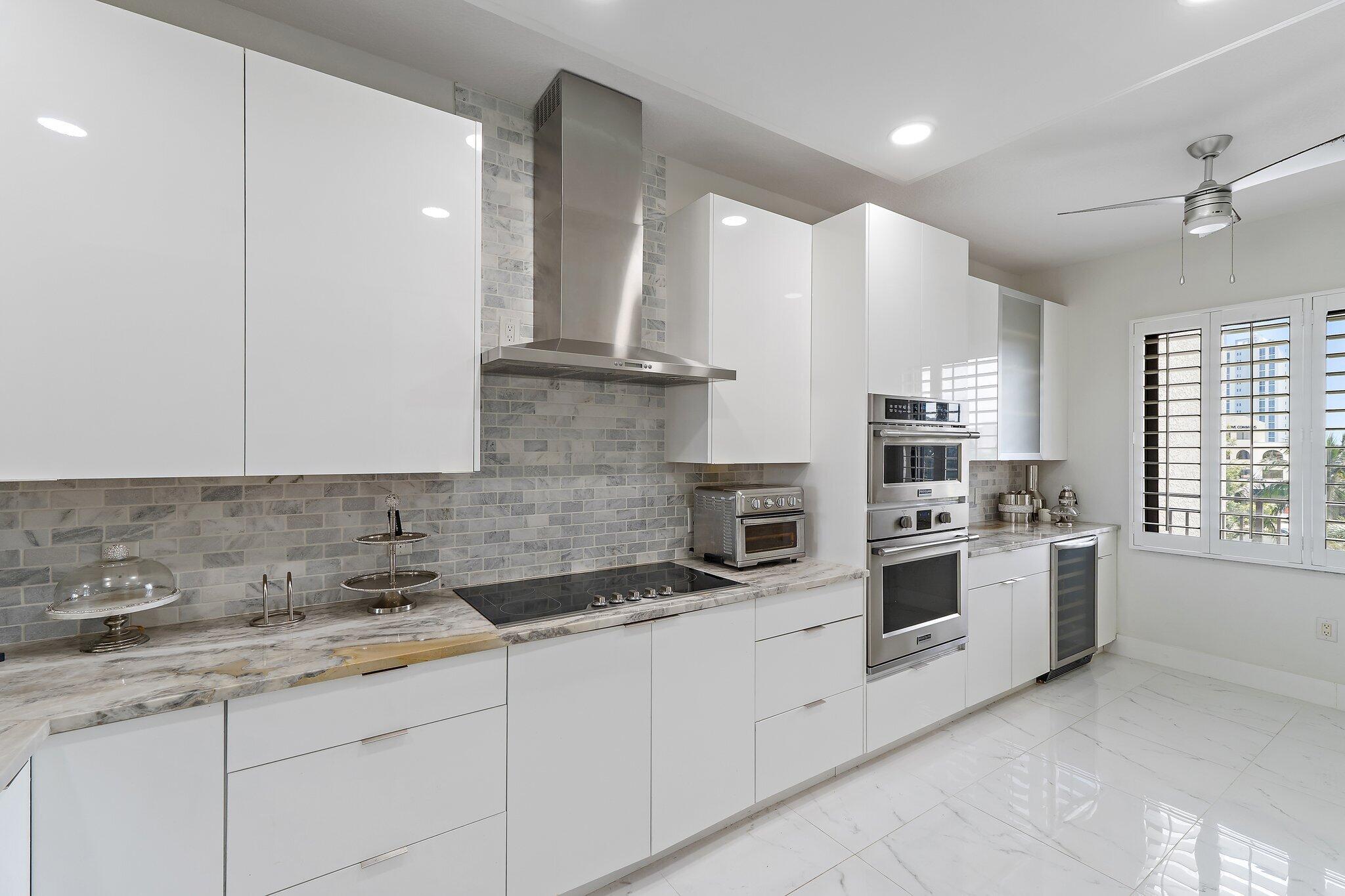 Modern white kitchen with marble counters, gray tile backsplash, stainless appliances, and shuttered windows.