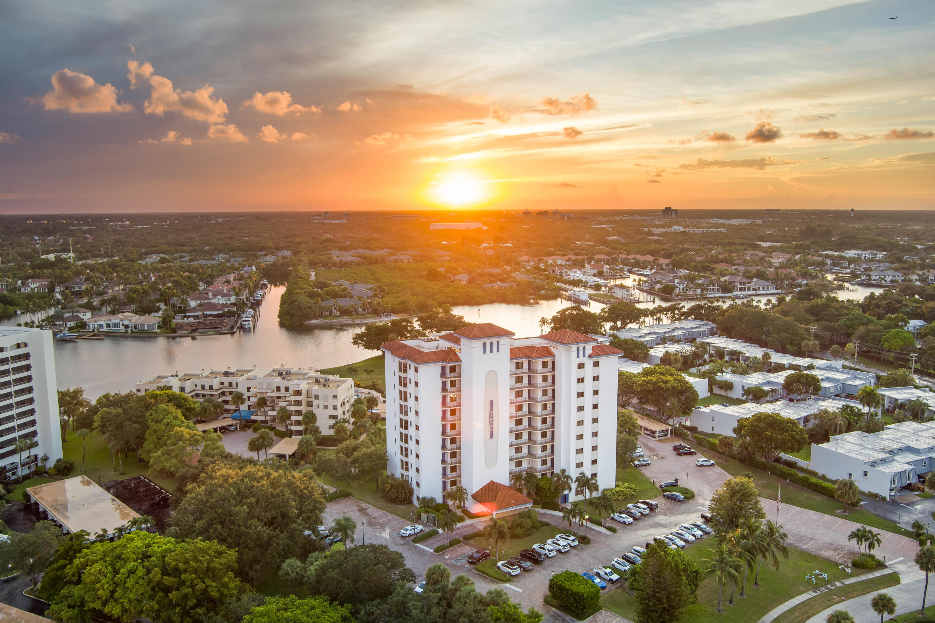 A white condominium tower beside a canal, surrounded by homes and palm trees at sunset.