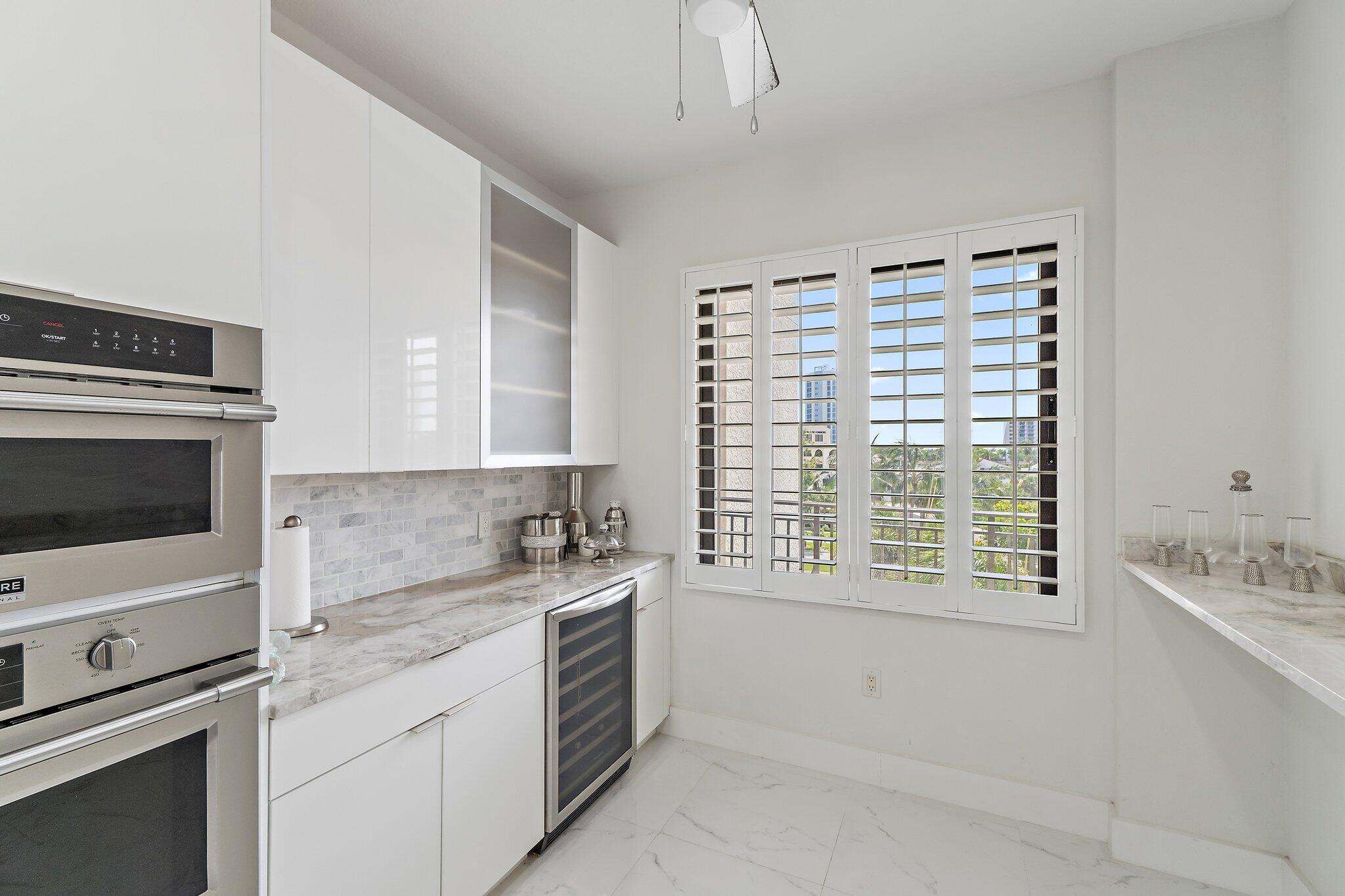 Modern kitchen with white cabinetry, marble counters, double ovens, and shuttered windows overlooking palms.