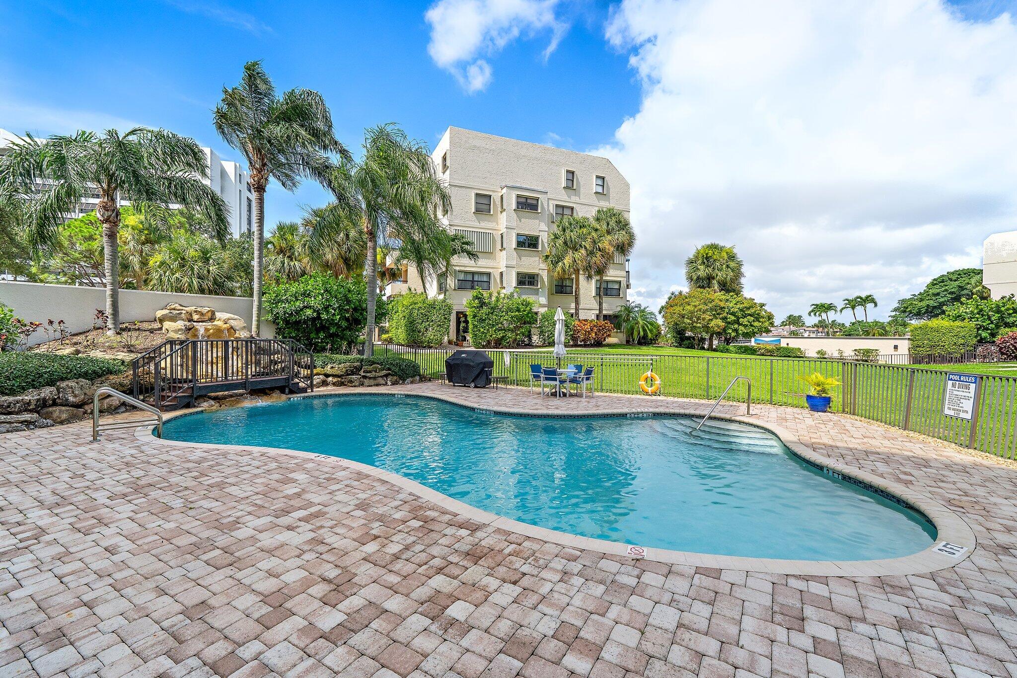 Community pool beside a condominium building, with a curved deck, palm trees, and a small footbridge.