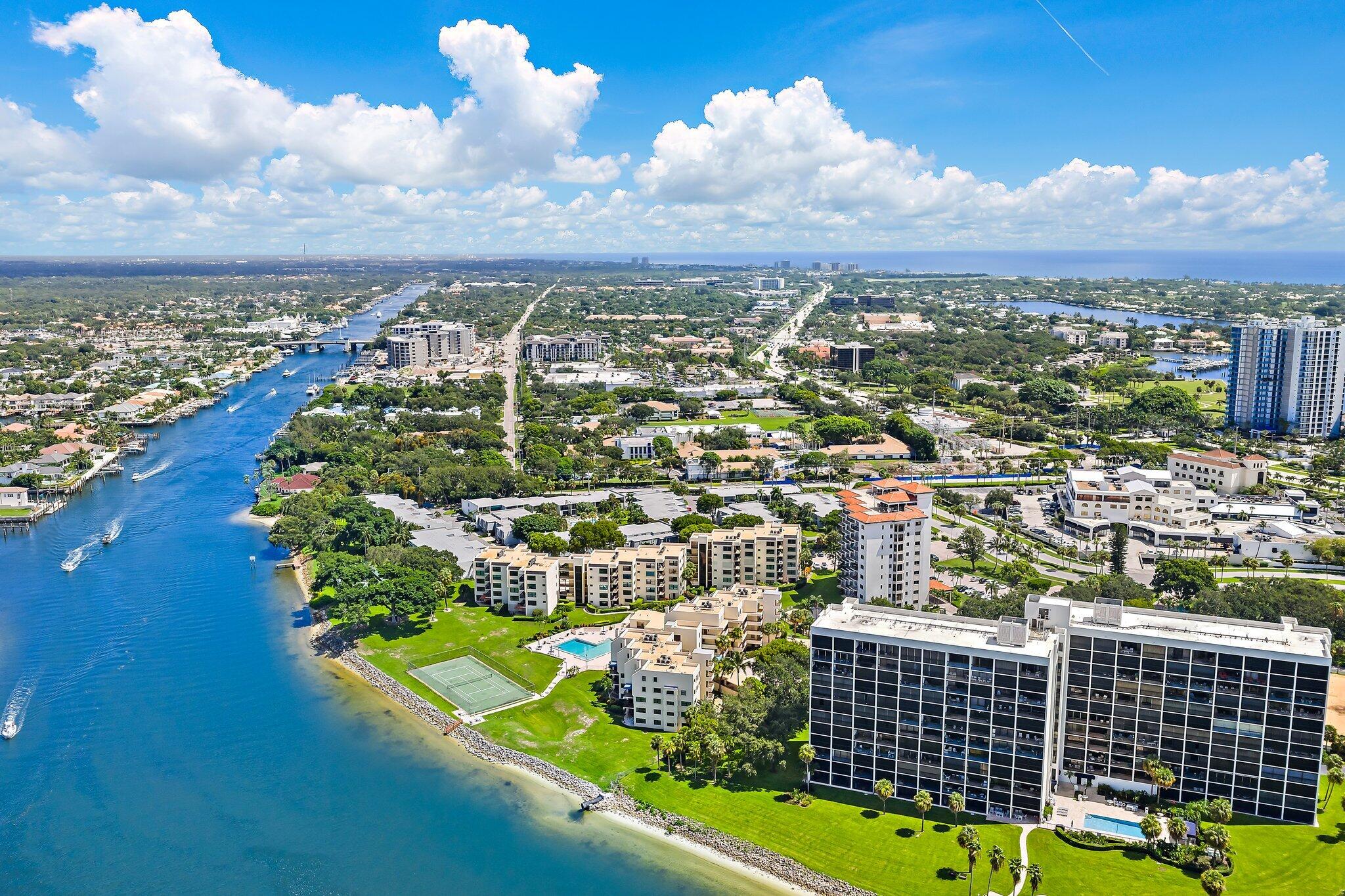 Aerial view of waterfront condominiums beside a broad waterway, with boats, a pool, and tennis court.