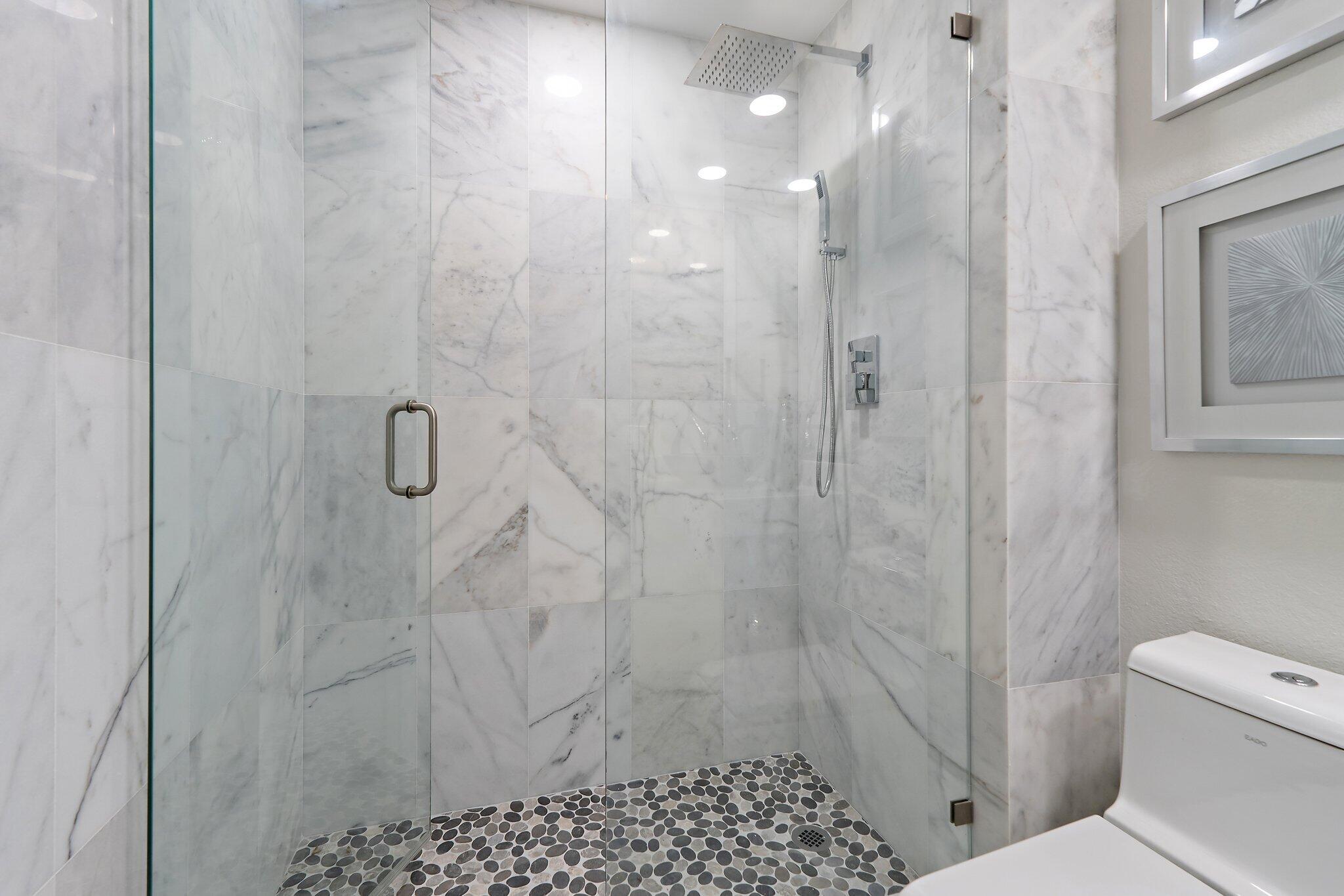 Glass-enclosed walk-in shower with marble-look walls, pebble floor, and rain showerhead.
