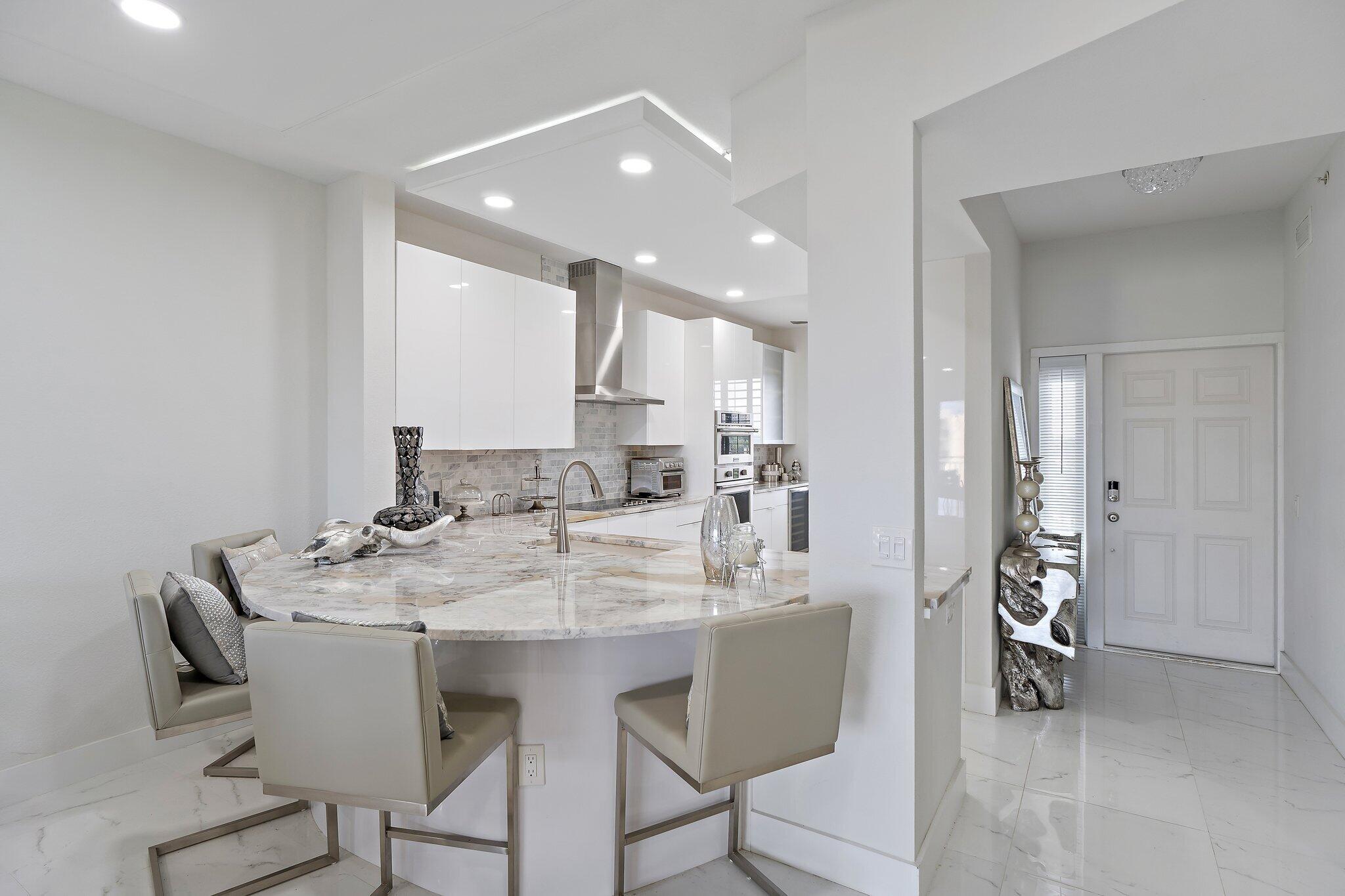 Modern white kitchen with a marble waterfall island, upholstered bar chairs, and stainless steel appliances.