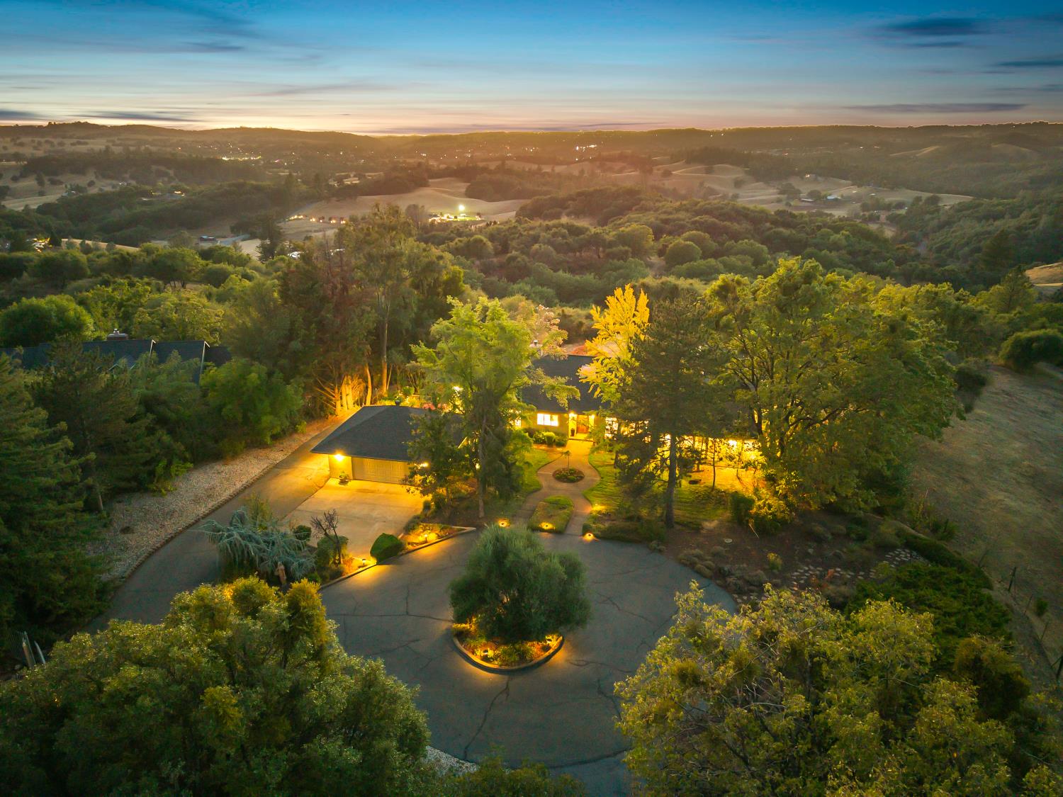 Aerial view of a tree-surrounded home and landscaped grounds at dusk, with rolling hills beyond.