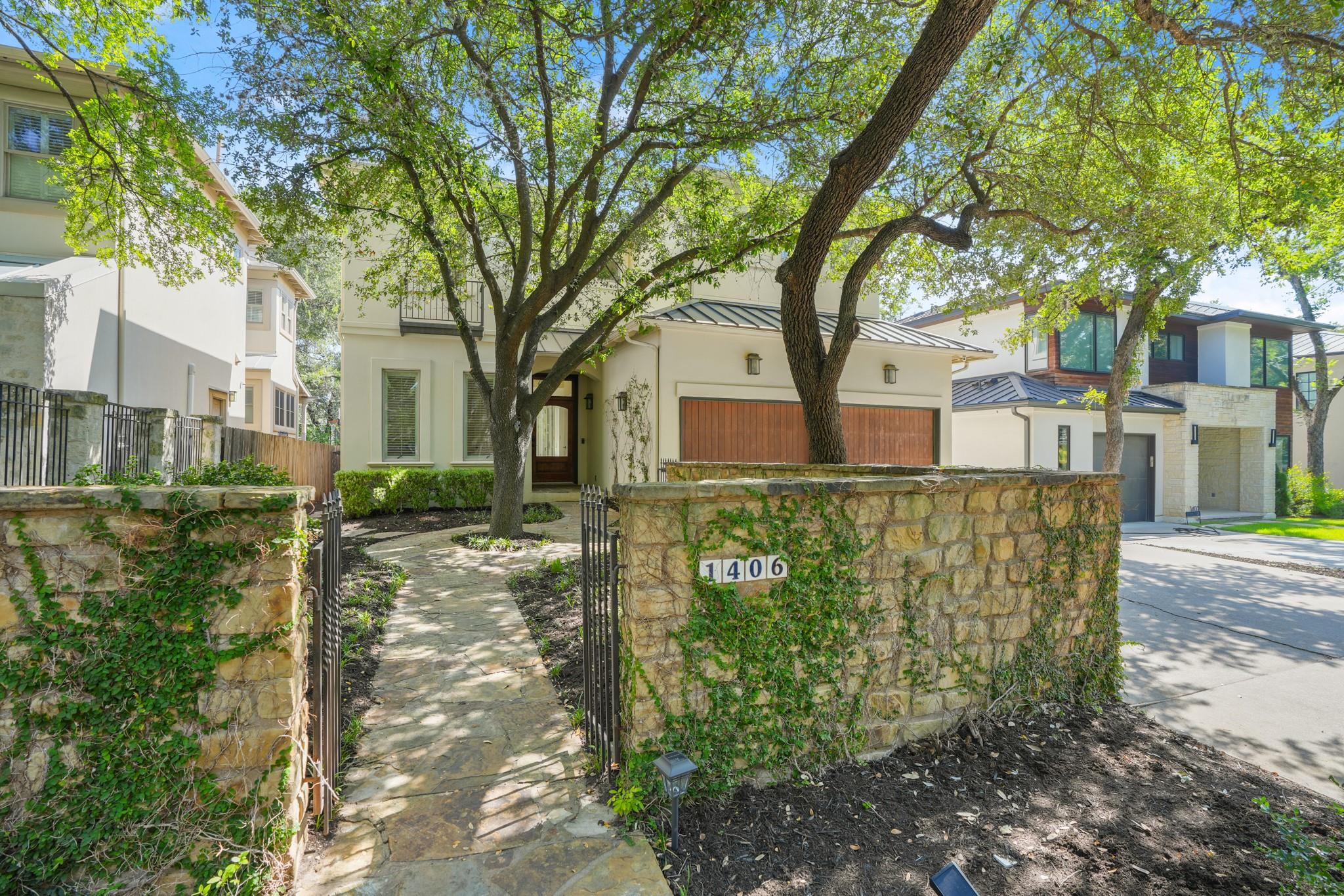 Cream-colored home with wooden garage doors, shaded by mature trees behind an ivy-covered stone wall.