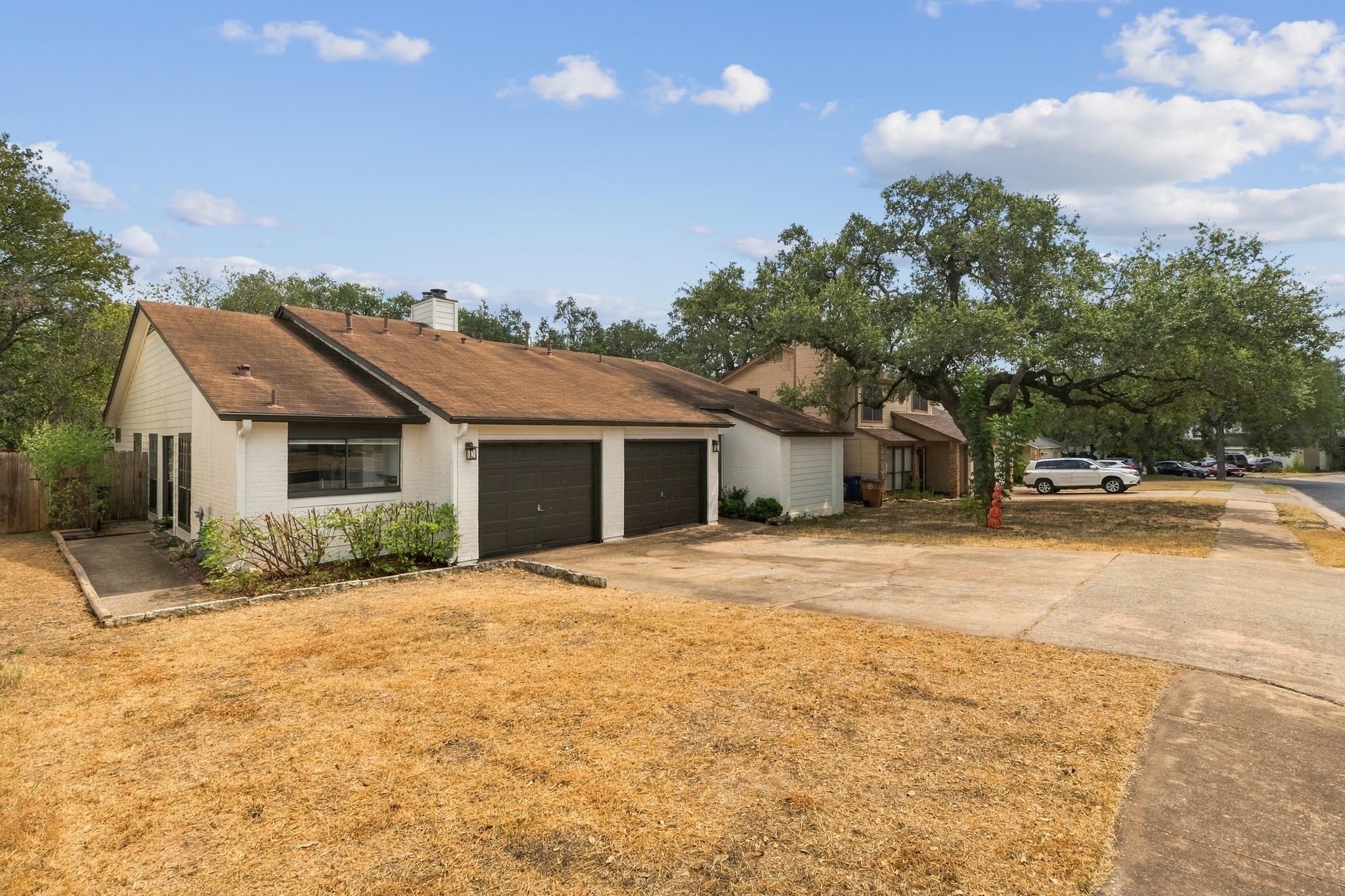 Single-story brick house with two dark garage doors, a broad driveway, and a large oak tree.